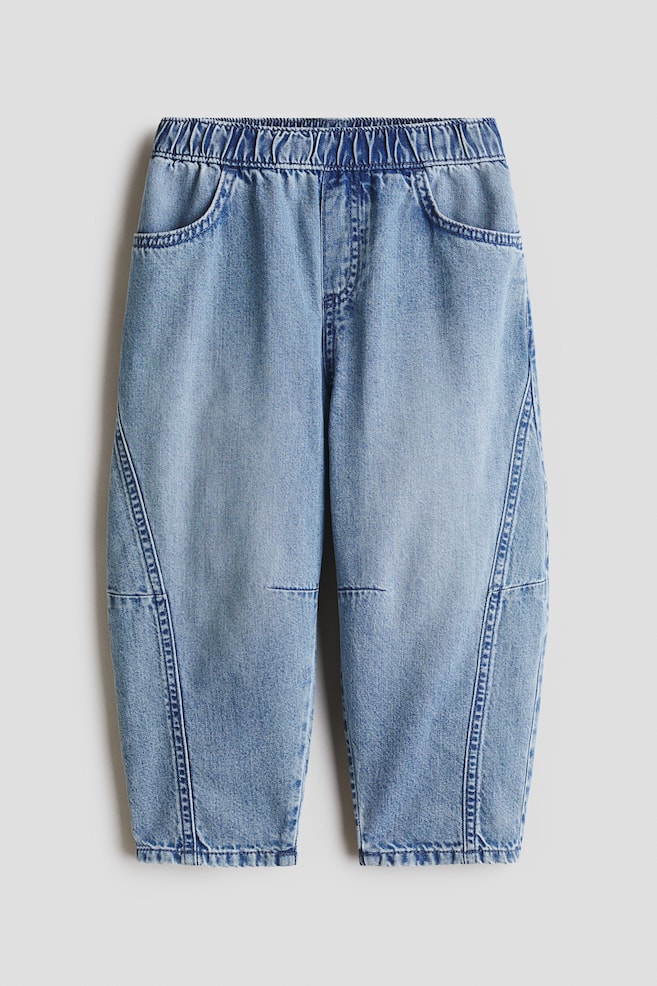 Light blue denim pants are showcasing a front view, designed with an elasticated waistband, two curved front pockets, and paneling crafted by diagonal seams on the outer legs and horizontal seams across the knees. - 1