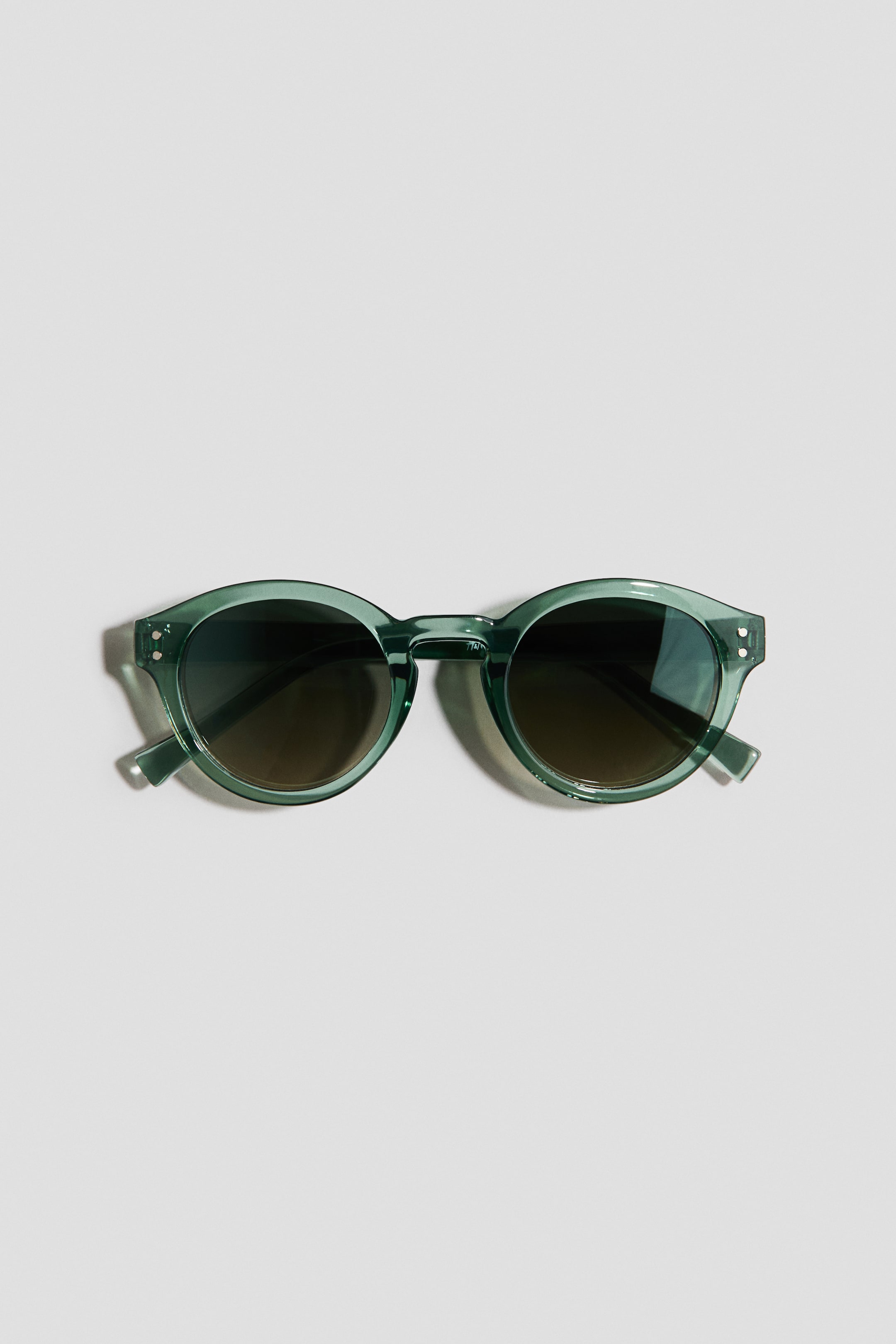 Ampliar la imagen: Las gafas de sol presentan un marco de plástico translúcido en color verde azulado y patillas a juego, con pequeños remaches plateados en las esquinas exteriores, sosteniendo lentes redondas de plástico con tinte oscuro.