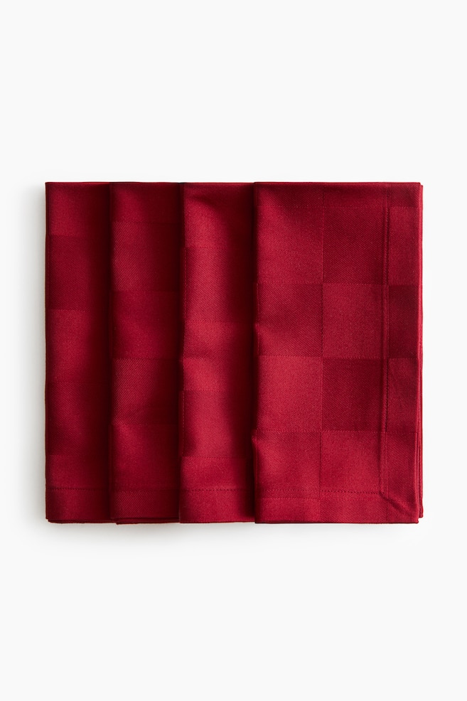 A set of four red fabric napkins lies in a slightly overlapping row, facing forwards, displaying a subtle woven chequerboard pattern. The folded napkins showcase stitched hems along their edges. - 1