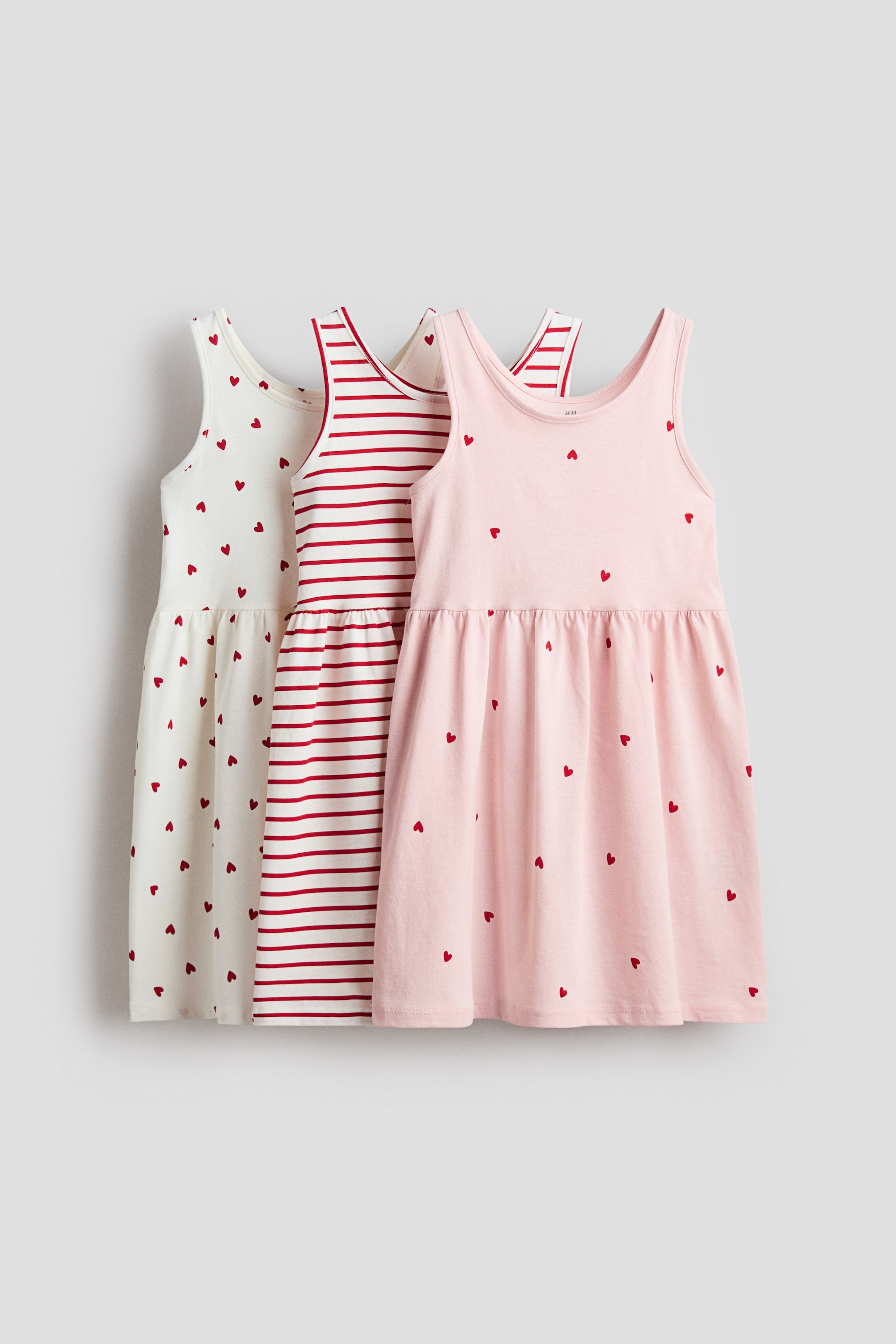 Größeres Bild anzeigen: Drei ärmellose Kleider zeigen ihre Vorderansicht, gefertigt aus weichem, bedrucktem Baumwolljersey. Jedes Kleid verfügt über einen runden Halsausschnitt, Raffungen an der Taille mit einem leicht ausgestellten Rock und ist entweder in Off-White mit roten Herzen, Weiß mit roten horizontalen Streifen oder Hellrosa mit roten Herzen gemustert.
