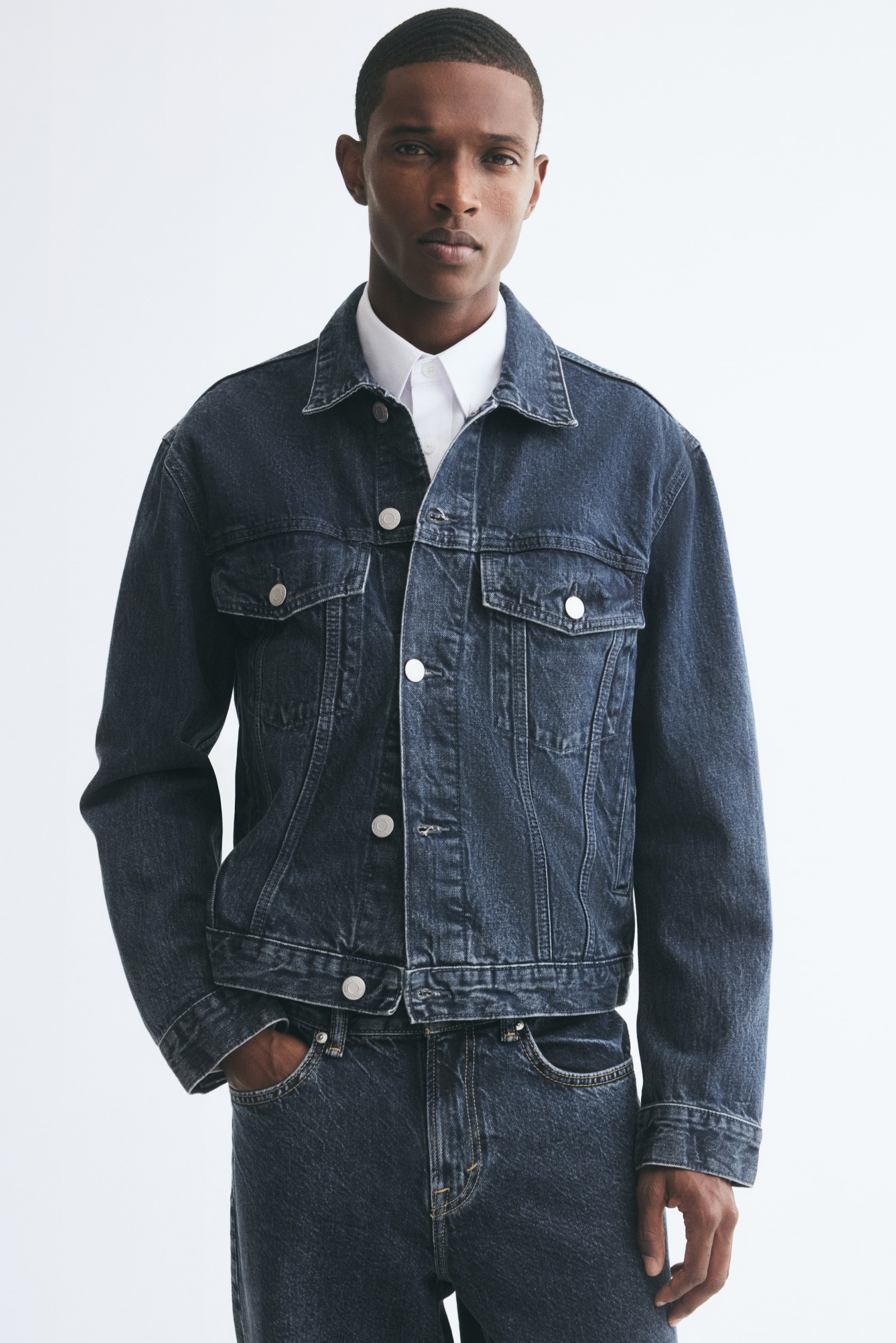 View larger image: The model is wearing a dark-wash denim jacket featuring a pointed collar, metal button closures down the front, and buttoned flap chest pockets. The jacket is layered over a white collared shirt and styled with matching denim jeans.