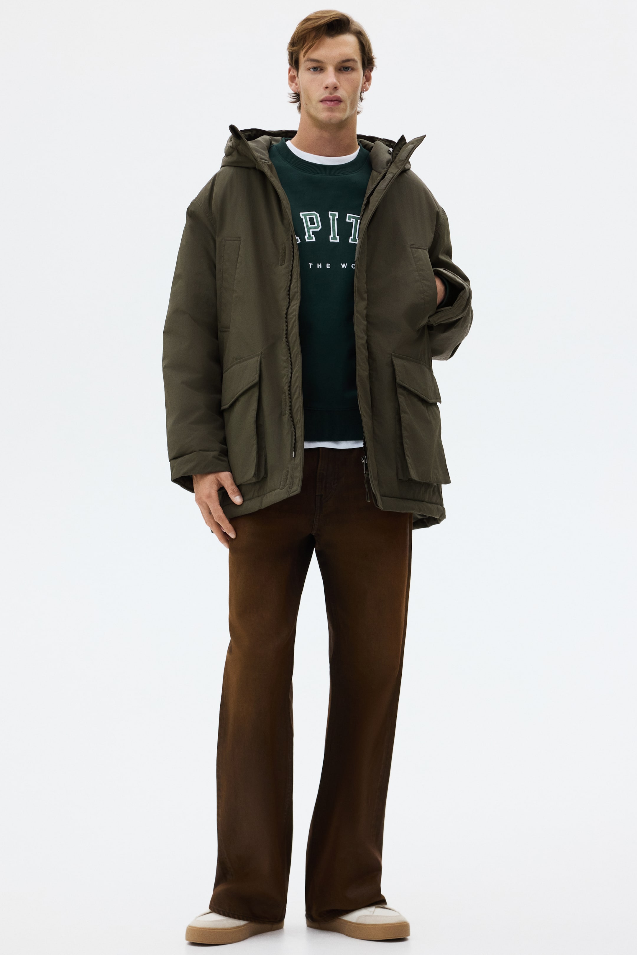 Ampliar la imagen: Una parka acolchada de color verde oliva está siendo usada por el modelo, con una capucha con cordón, un cuello de pie y bolsillos frontales de parche con solapas. Su cierre frontal con tapeta contra el viento está parcialmente abierto, y el modelo está colocando una mano en un bolsillo para calentar las manos, combinando la parka sobre una sudadera verde oscuro.