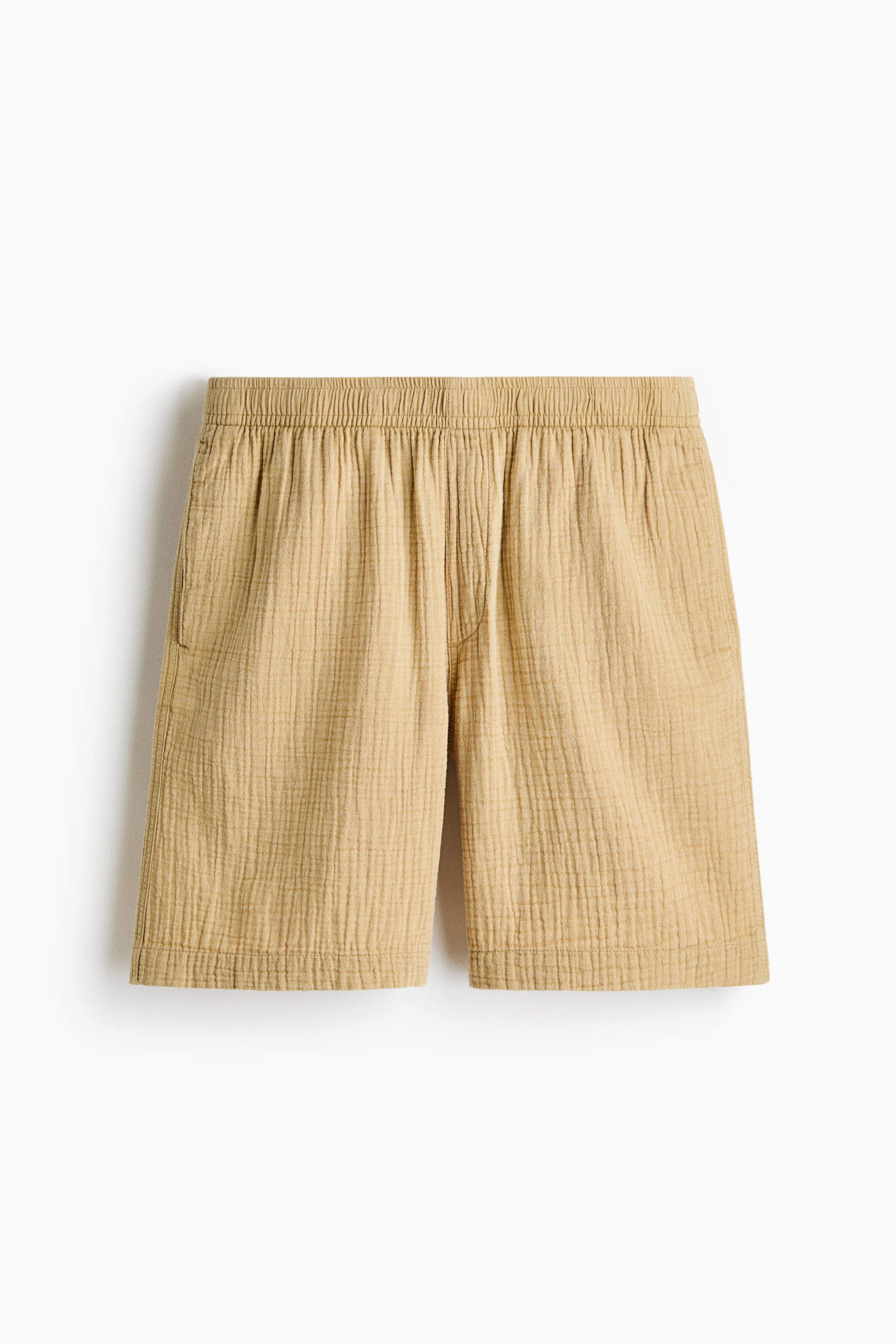View larger image: Tan cotton muslin shorts showcase a relaxed fit and crinkled texture at the front. The waist features covered elastication, with a subtle side pocket opening visible on the left.