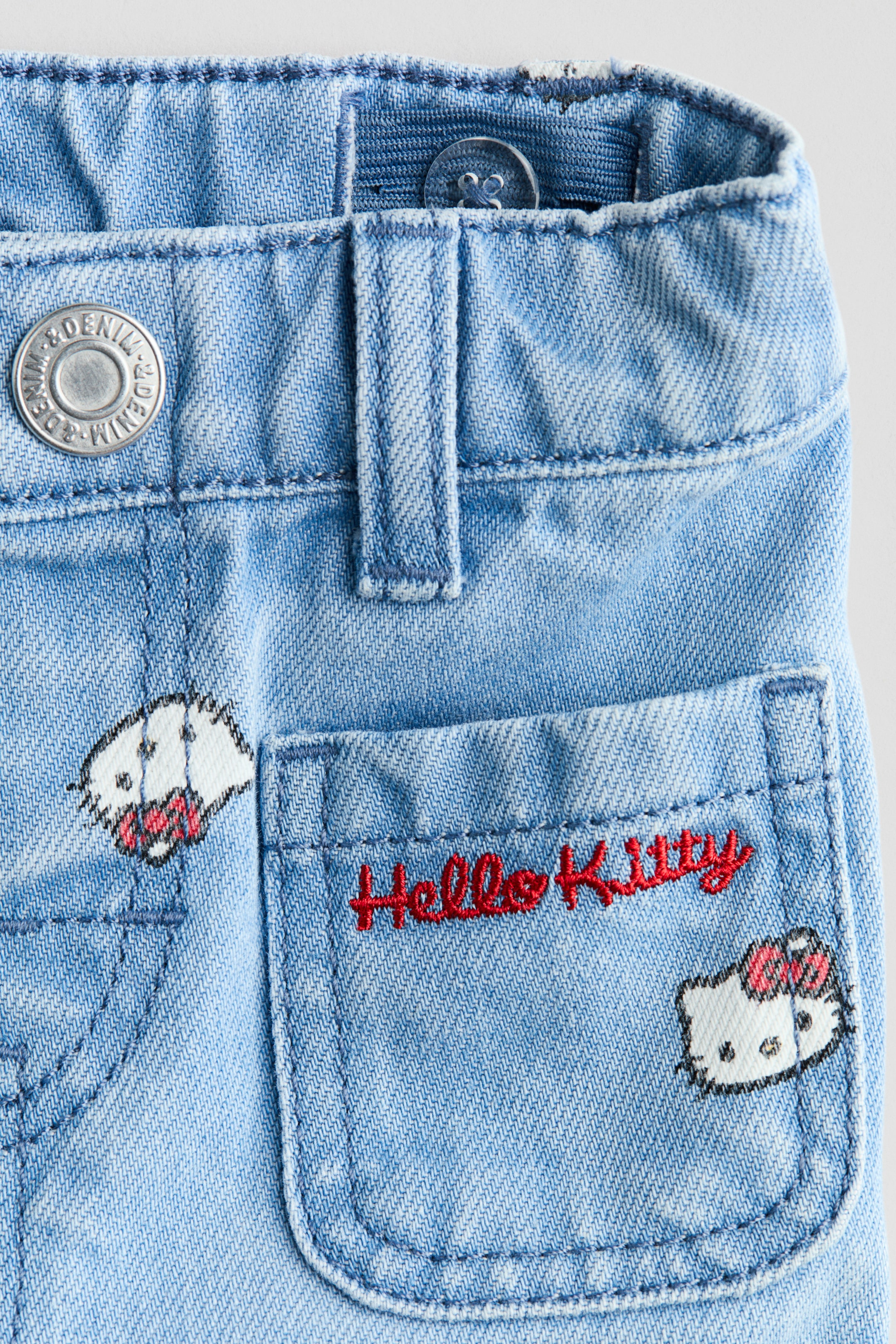 Ampliar la imagen: Pantalones de mezclilla azul claro presentan una pretina con trabillas para cinturón, una banda elástica ajustable interna y un botón de metal. Un bolsillo patch en el frente muestra un bordado con la inscripción roja "Hello Kitty" y dos caras de Hello Kitty bordadas con lazos rosas.