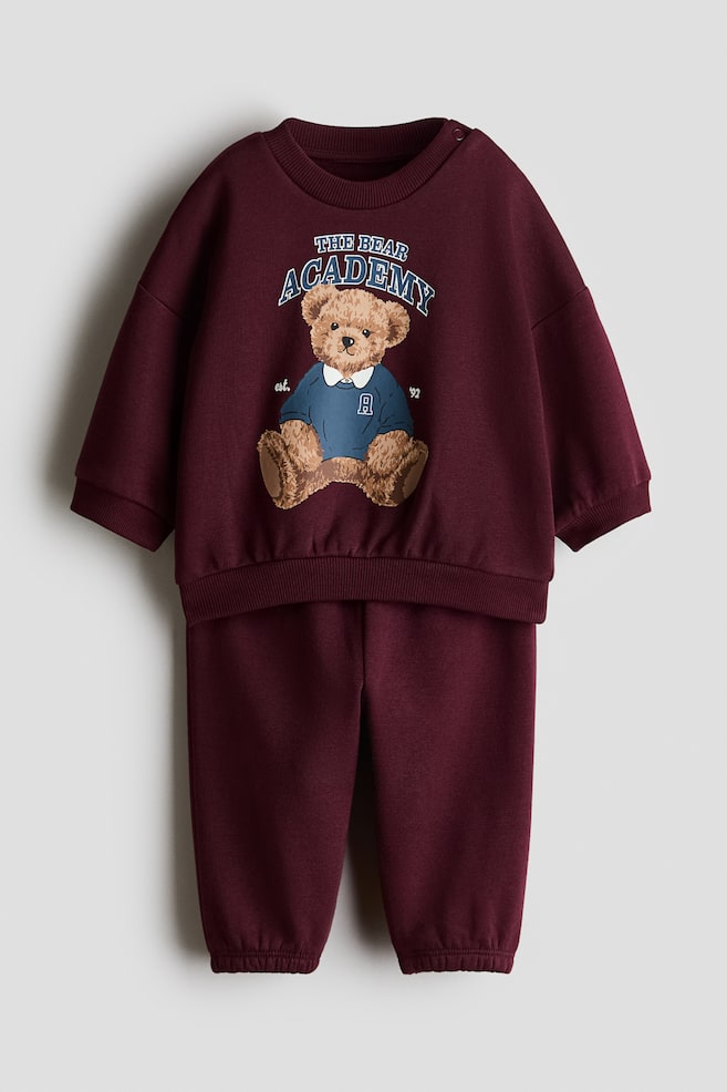 A 2-piece printed sweat fabric set in burgundy is showcasing a front view of a long-sleeved sweatshirt and matching sweatpants. The sweatshirt features dropped shoulders, ribbed trims, and a central graphic of a teddy bear wearing a blue sweater beneath the text "THE BEAR ACADEMY". The matching sweatpants complete the set, with a relaxed fit and elasticised ankle cuffs. - 1