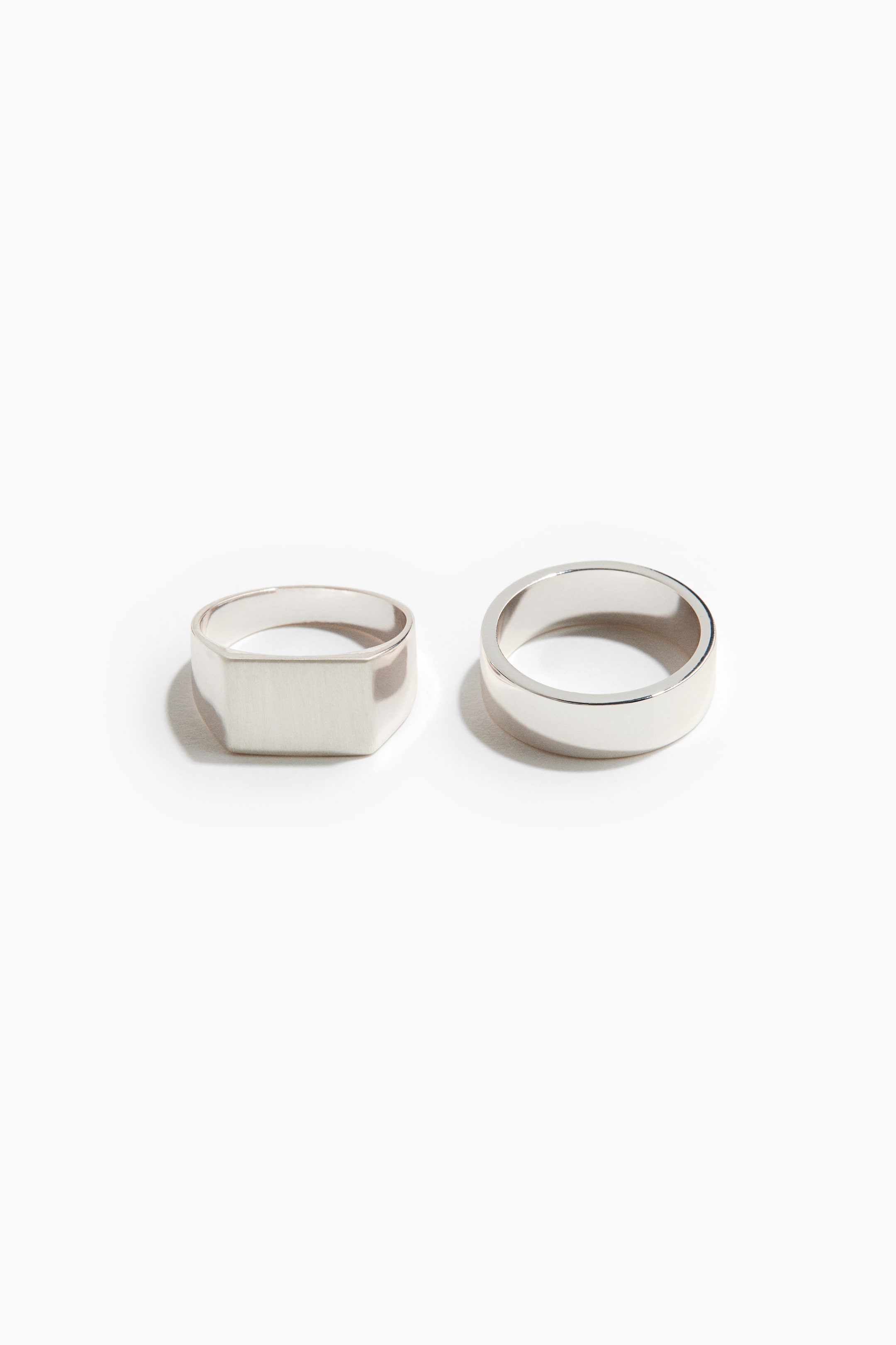 View larger image: A pair of silver-toned rings is placed side by side, with the left ring featuring a flat, rectangular signet face with a brushed finish and the right ring showcasing a classic wide band with a polished finish.