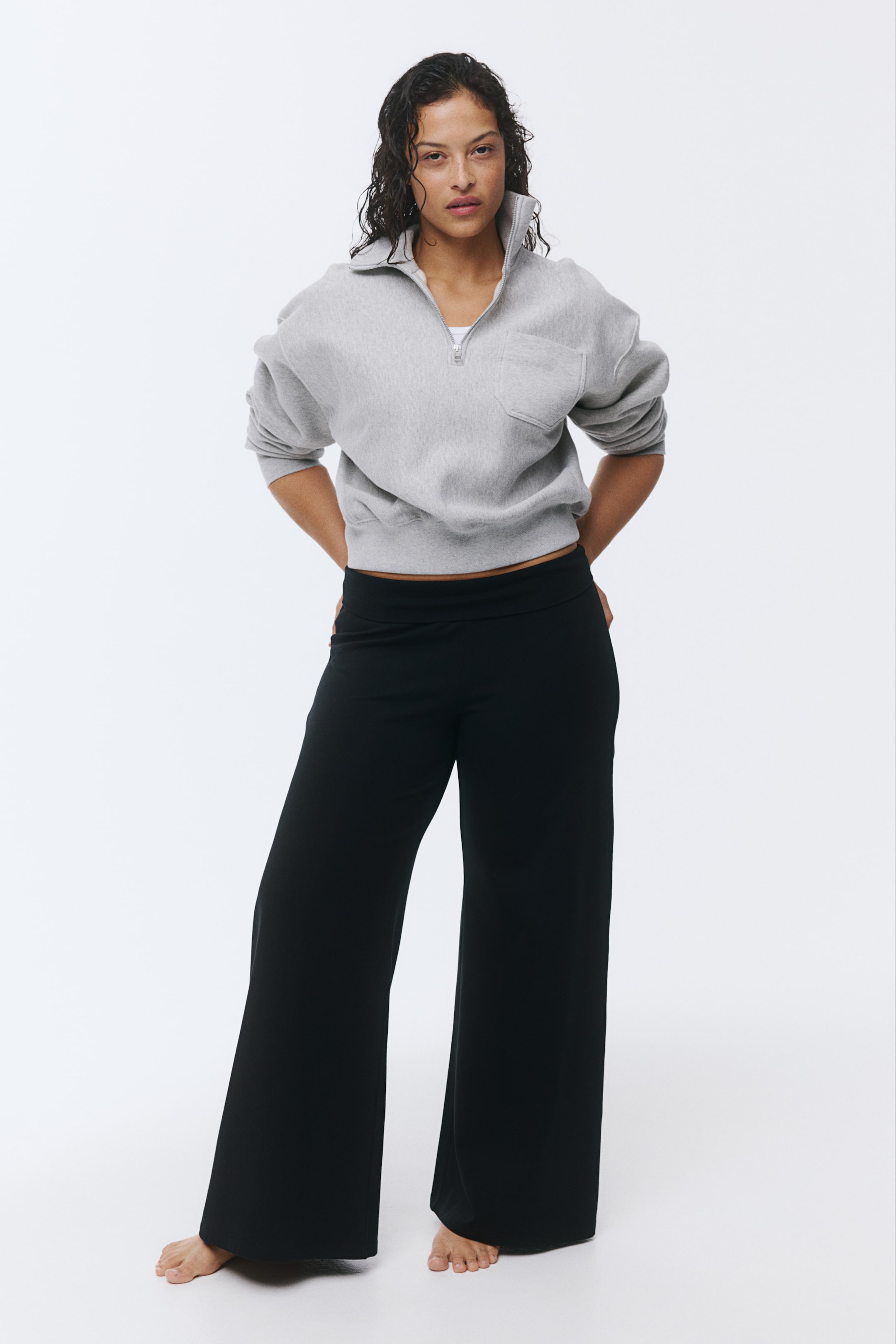 View larger image: A grey quarter-zip sweatshirt is being presented, featuring a stand-up collar, a silver-toned zipper revealing a white undershirt, and a patch pocket on the left chest. This cropped sweatshirt is being paired with black wide-leg pants, which are showing a wide waistband and a flowing silhouette.