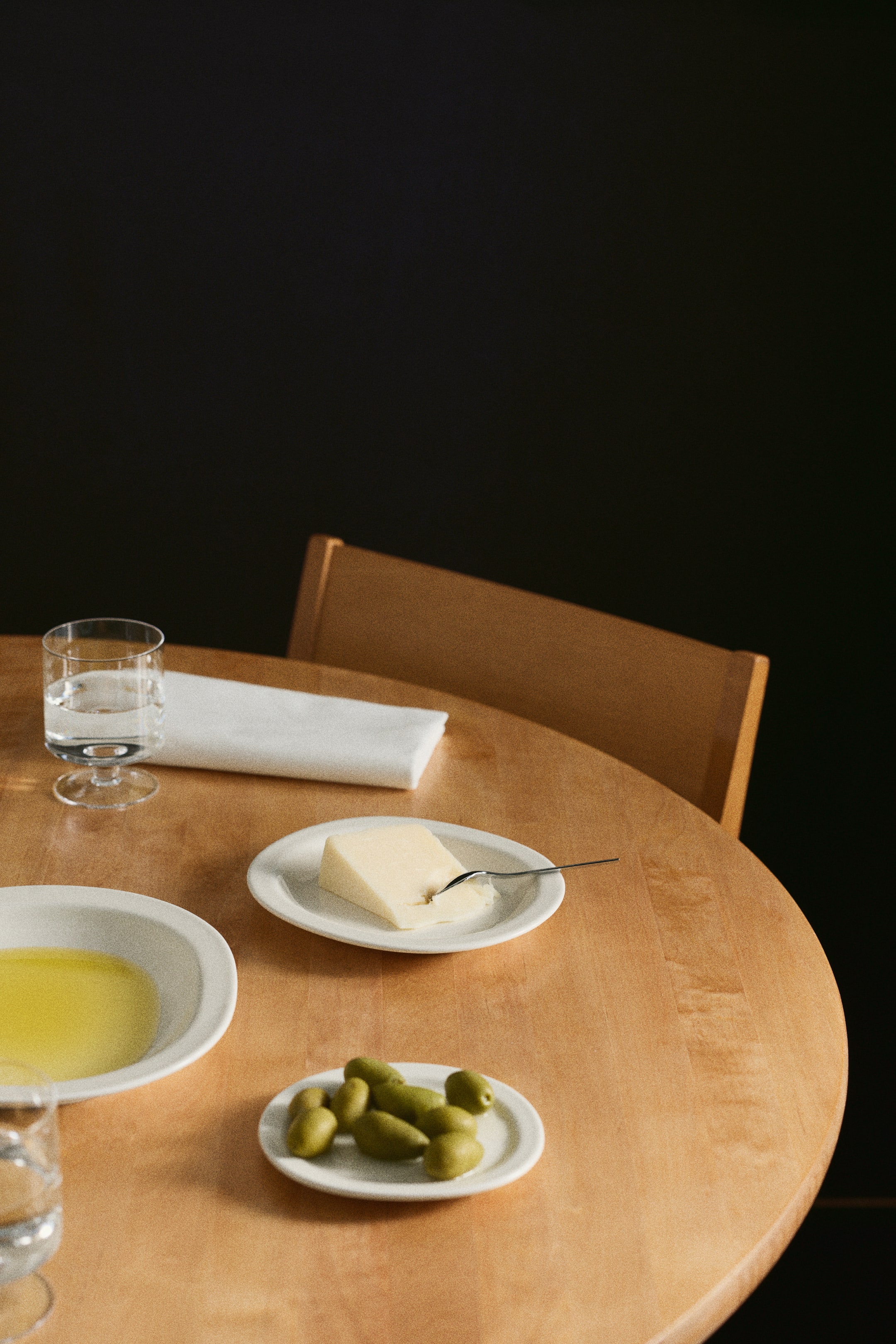 View larger image: Three off-white porcelain plates with a minimalist design and classic rim display food items. A deep-rimmed plate holds olive oil, a medium plate presents a wedge of cheese with a fork, and a smaller plate holds green olives.