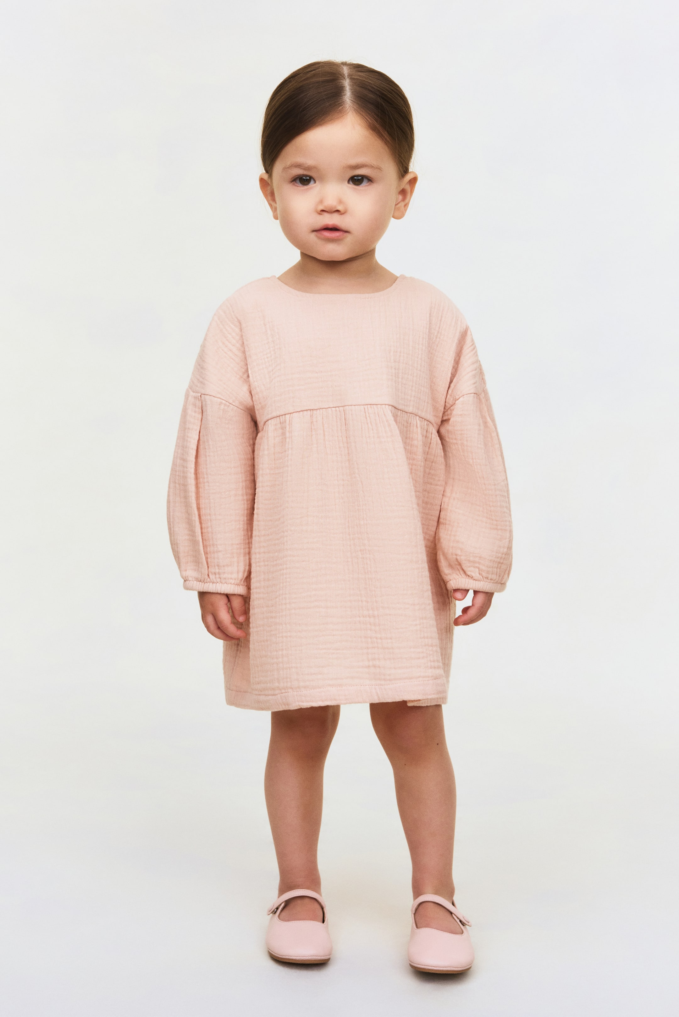 View larger image: A light pink smock dress features a crinkled texture, a round neckline, and long, voluminous sleeves with gathered cuffs. The dress has a gathered seam at the empire waist that flows into a relaxed A-line skirt.