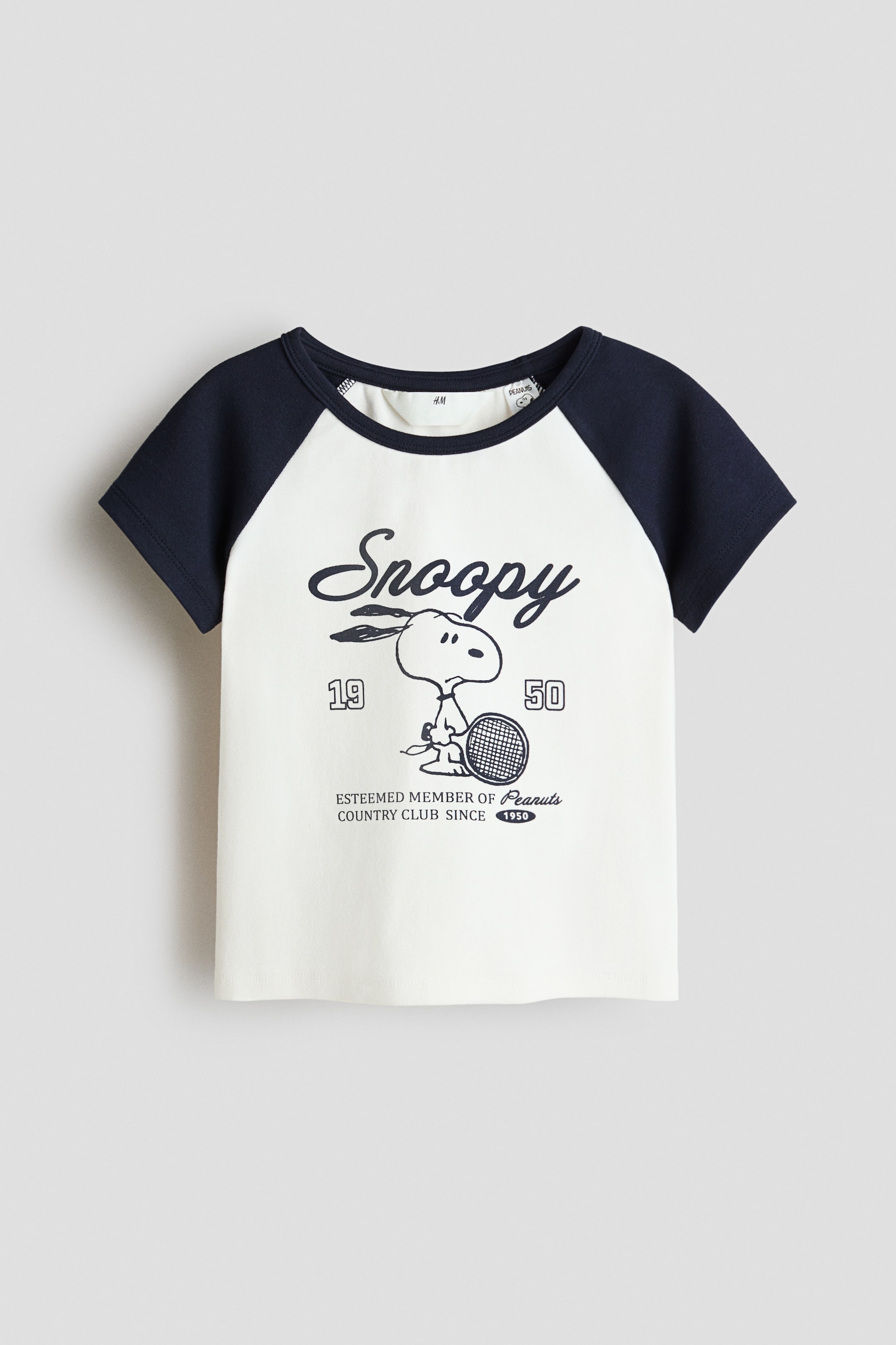 View larger image: The front of this short-sleeved top features a cream-coloured body with navy blue raglan sleeves and a matching round neckline. The front showcases a print motif of the word "Snoopy" in script above an illustration of Snoopy holding a tennis racket, accompanied by the text "ESTEEMED MEMBER OF Peanuts COUNTRY CLUB SINCE 1950".