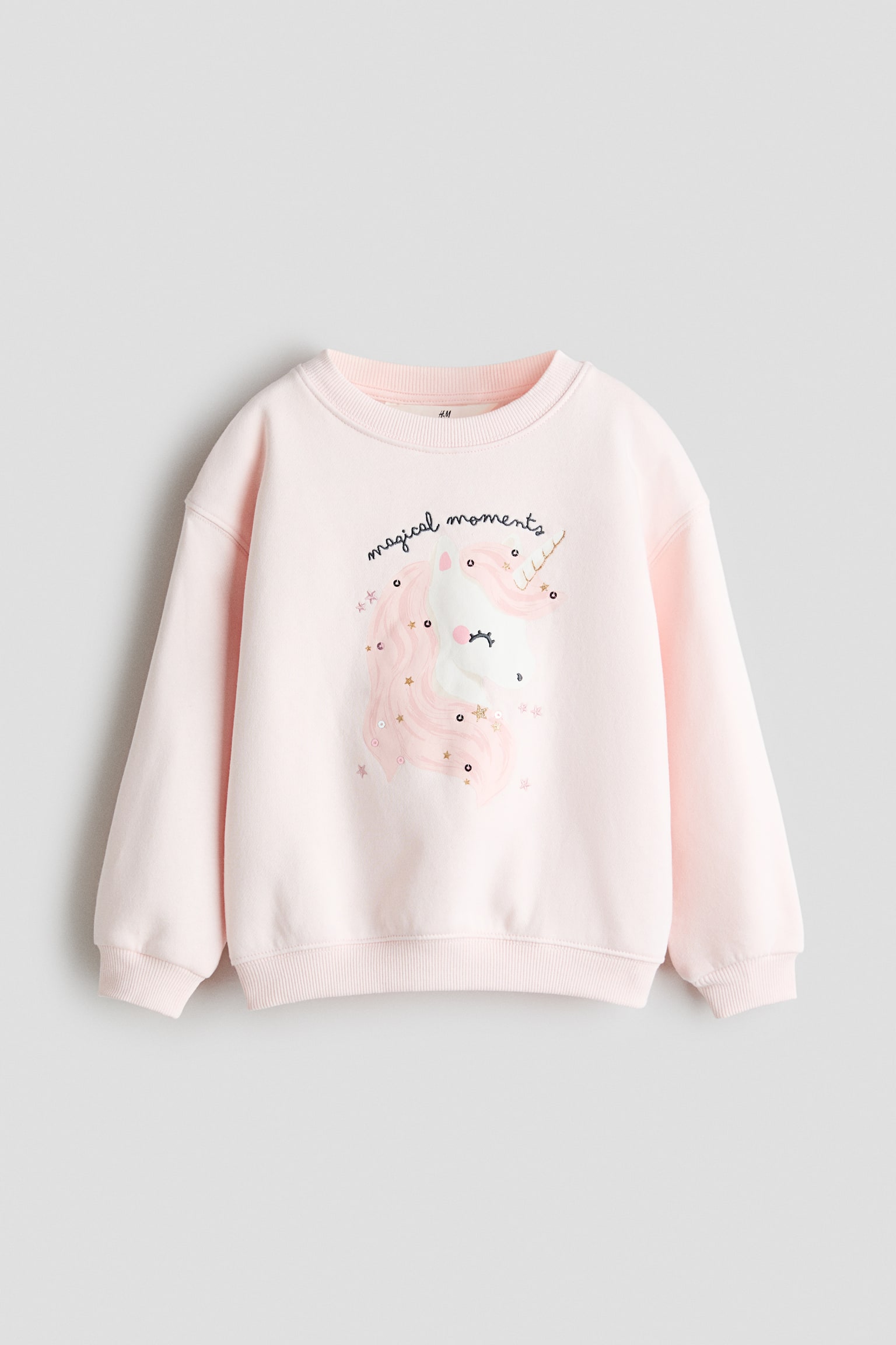 A light pink sweatshirt is shown from the front, featuring a round, rib-trimmed neckline, dropped shoulders, and ribbed cuffs and hem. The front displays a graphic of a unicorn with a pink mane and gold horn beneath the cursive text "magical moments", with small star and sequin details. - 1