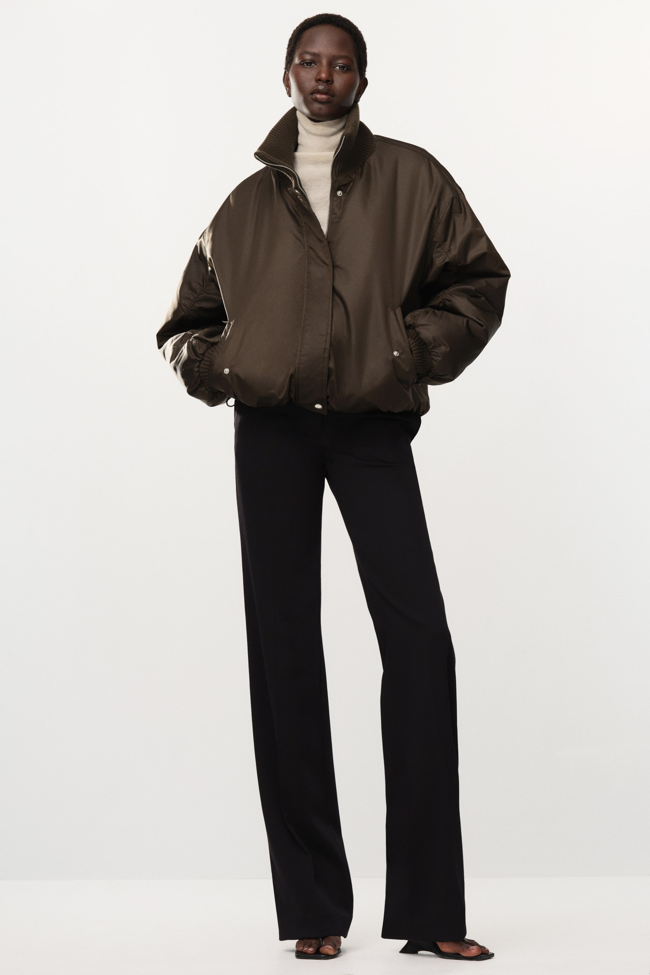 Größeres Bild ansehen: Eine oversized, gepolsterte braune Bomberjacke aus Nylon wird vom Model getragen, wobei die Hände in den Seitentaschen ruhen. Die Jacke verfügt über einen hohen, offenen Rippkragen und überschnittene Schultern und wird über einem cremefarbenen Rollkragenoberteil und einer schwarzen Wide-Leg-Hose getragen.