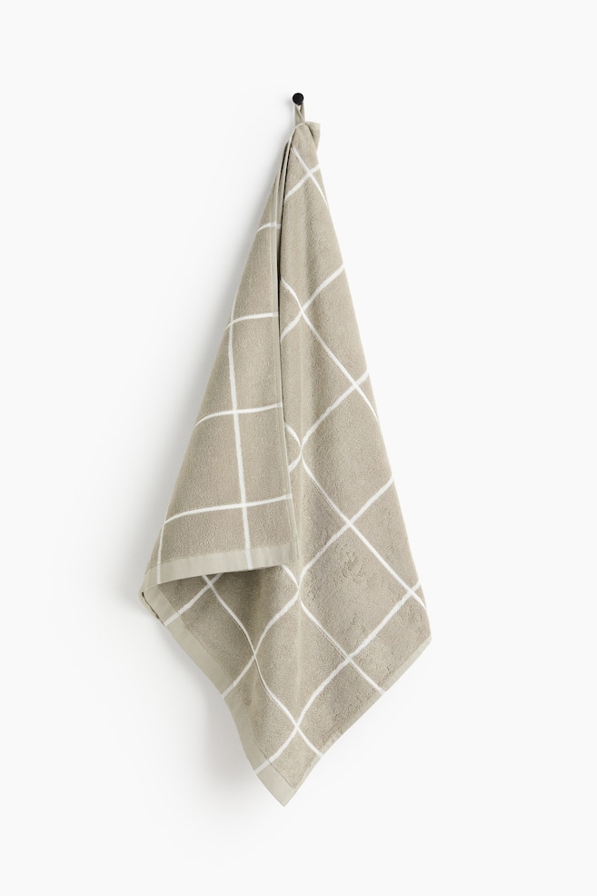 The towel is a light brown terry cloth, elegantly draped from a black hook. It features a white grid pattern across its surface and is neatly finished with hemmed edges. - 1