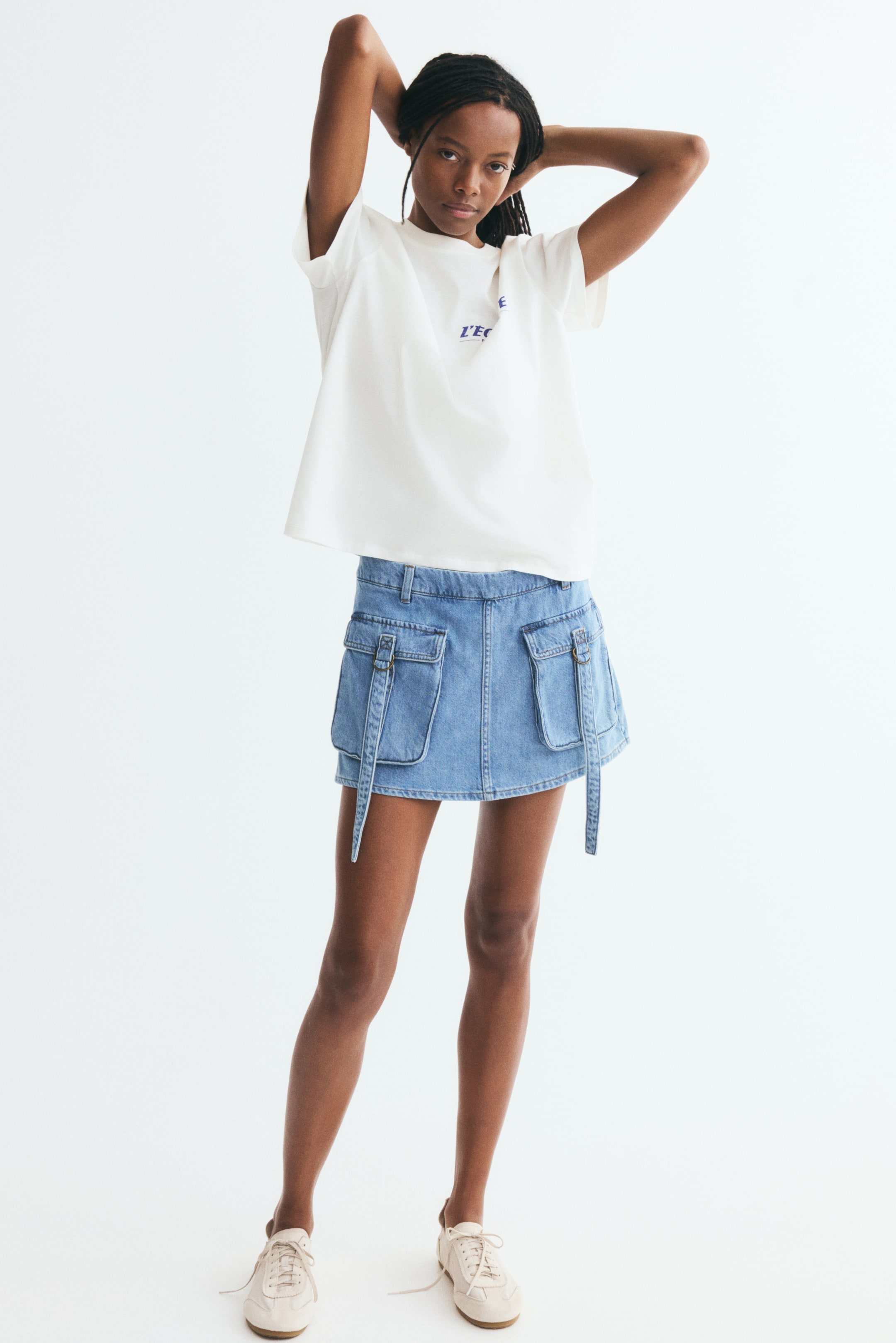 Xem ảnh lớn hơn: Một chiếc áo thun dáng rộng bằng jersey cotton màu trắng ngà đang tạo nên phom dáng thoải mái khi nhìn từ phía trước, với cổ tròn bo gân, vai xuôi và họa tiết chữ in màu xanh nhỏ trên ngực. Áo được thả rủ tự nhiên bên ngoài chân váy denim xanh nhạt, làm nổi bật kiểu dáng rộng rãi, phóng khoáng của sản phẩm.