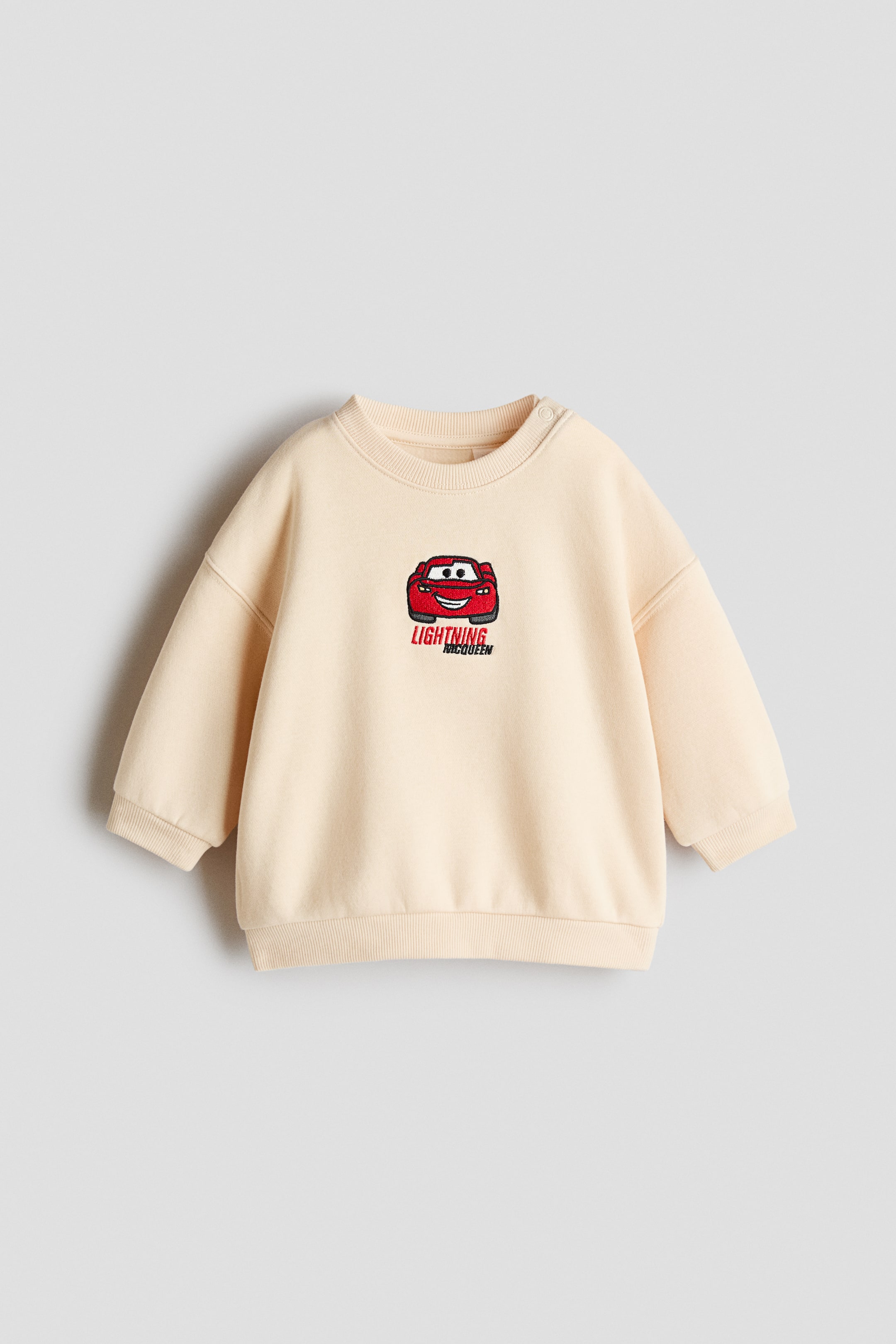 View larger image: The cream-colored sweatshirt is showing its FRONT. It is featuring an embroidered red car character with "Lightning McQueen" text below it, and it is displaying a ribbed crew neck, ribbed cuffs, a ribbed hem, and two snap buttons on the right shoulder.