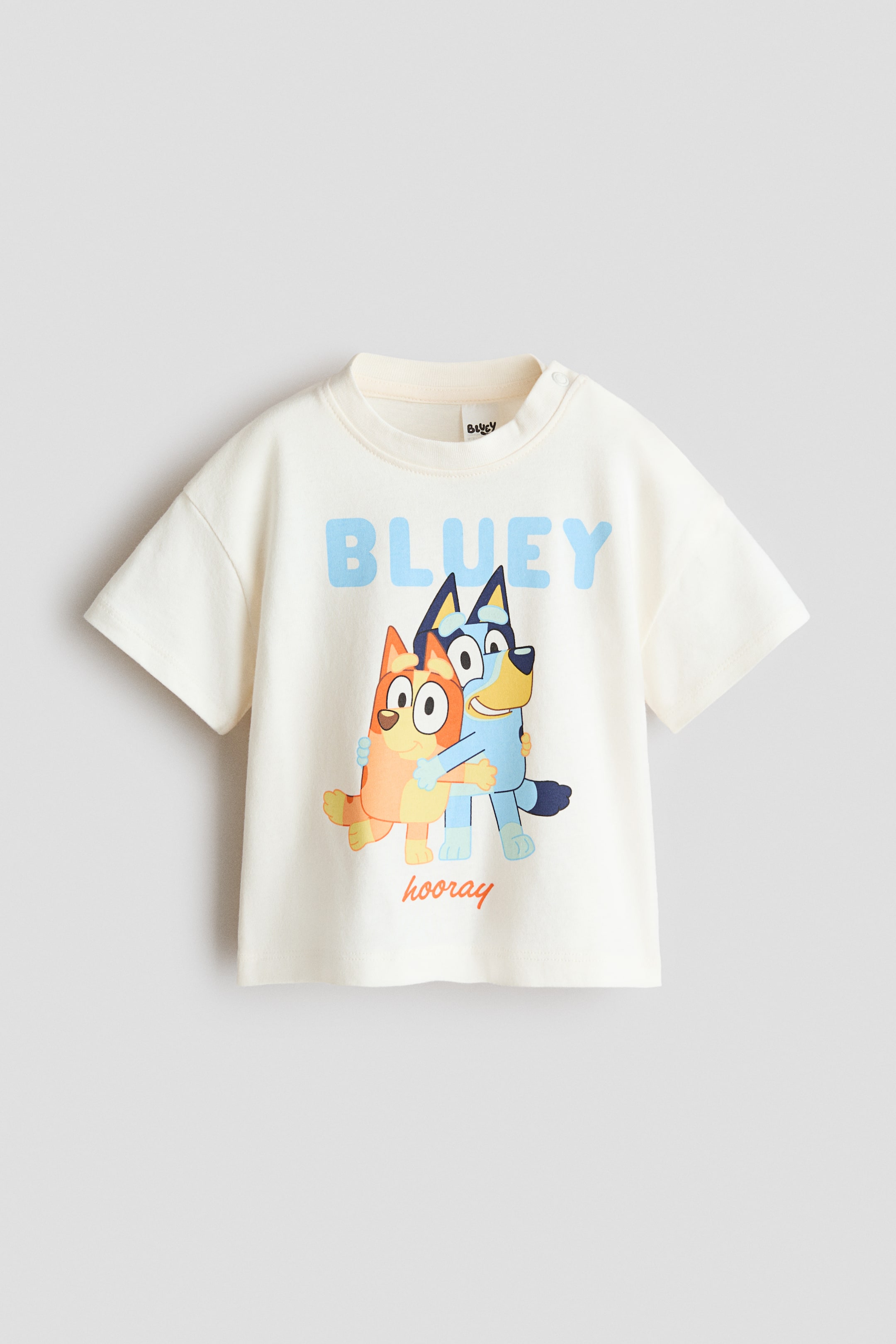 View larger image: The front of this printed cotton T-shirt is shown in an off-white hue. It showcases a light blue "BLUEY" graphic centered above two embracing cartoon dog characters, with "hooray" printed underneath. Two snap buttons are visible on the left shoulder.