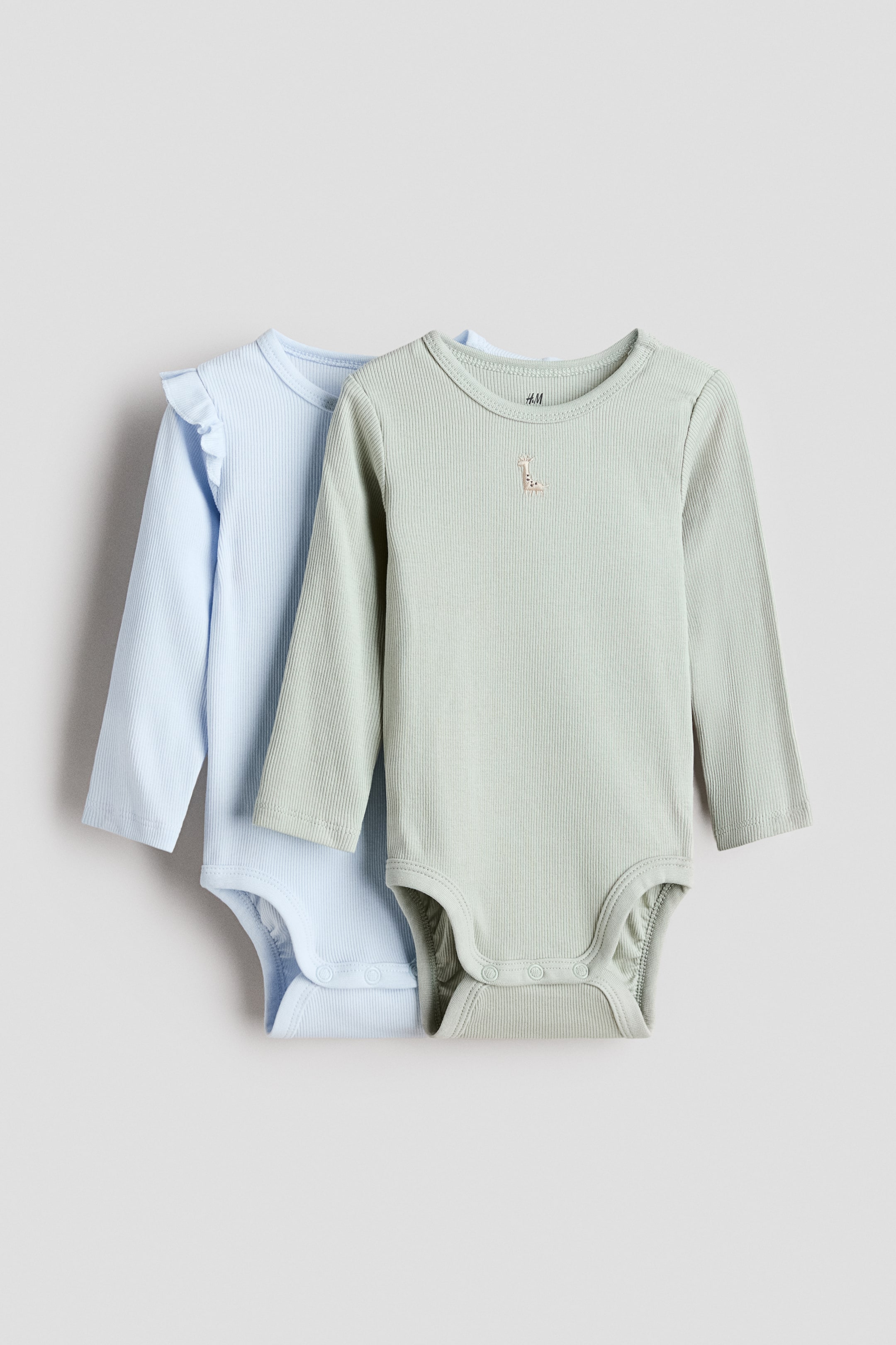 Größeres Bild anzeigen: Zwei langärmelige Baby-Body-Anzüge sind von vorne ausgestellt. Der hellgrüne Body vorne hat eine gerippte Struktur und eine kleine gestickte Giraffe auf der Brust. Der hellblaue Body dahinter hat ebenfalls eine gerippte Struktur und Rüschen an den Schultern, und beide Bodys haben Druckknöpfe im Schritt.