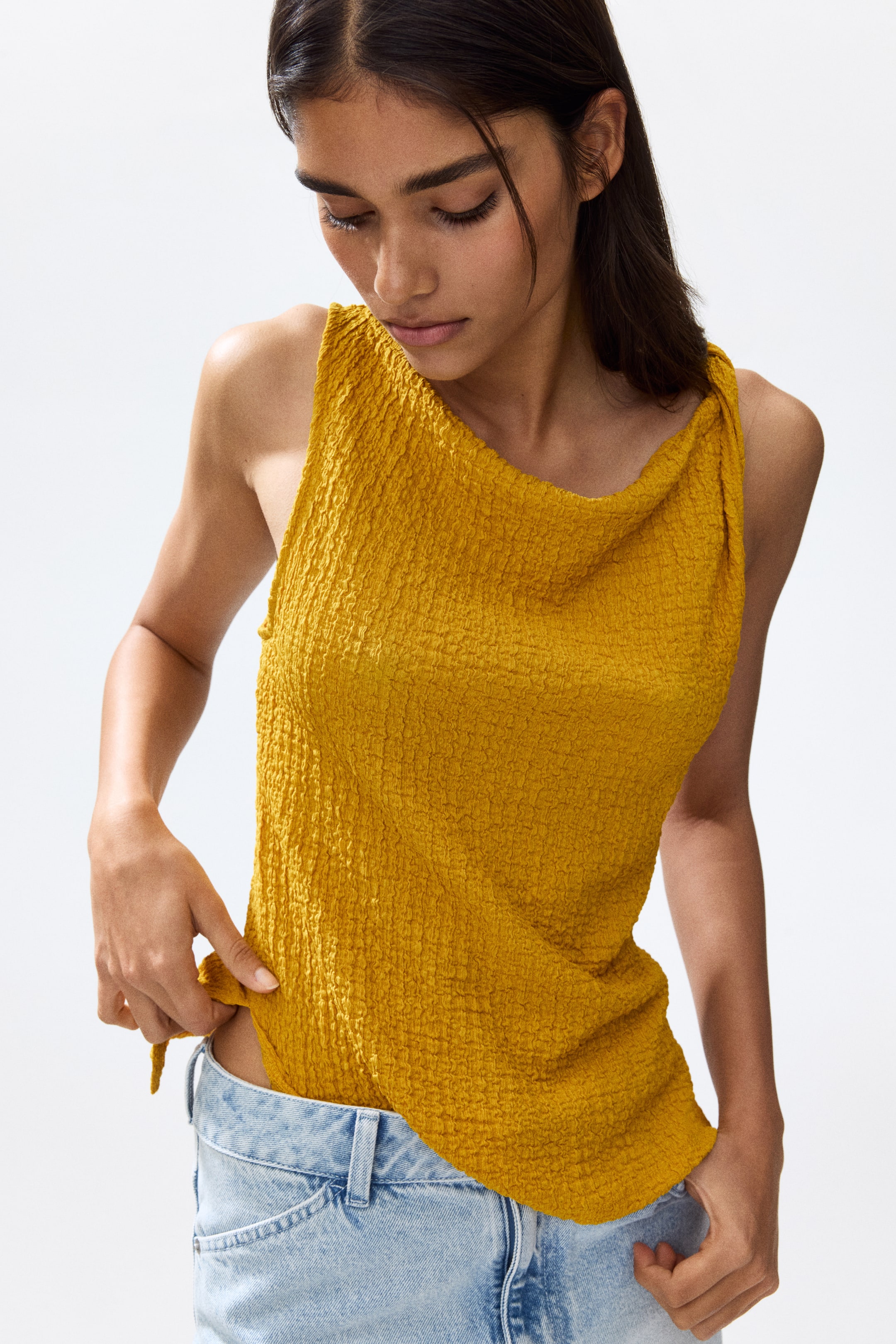 View larger image: The model is wearing a sleeveless, fitted top crafted from yellow crinkled jersey. It showcases an asymmetric neckline, a single twisted shoulder strap, and side slits at the hem. The model is adjusting the top, which is partially tucked into light-wash jeans.