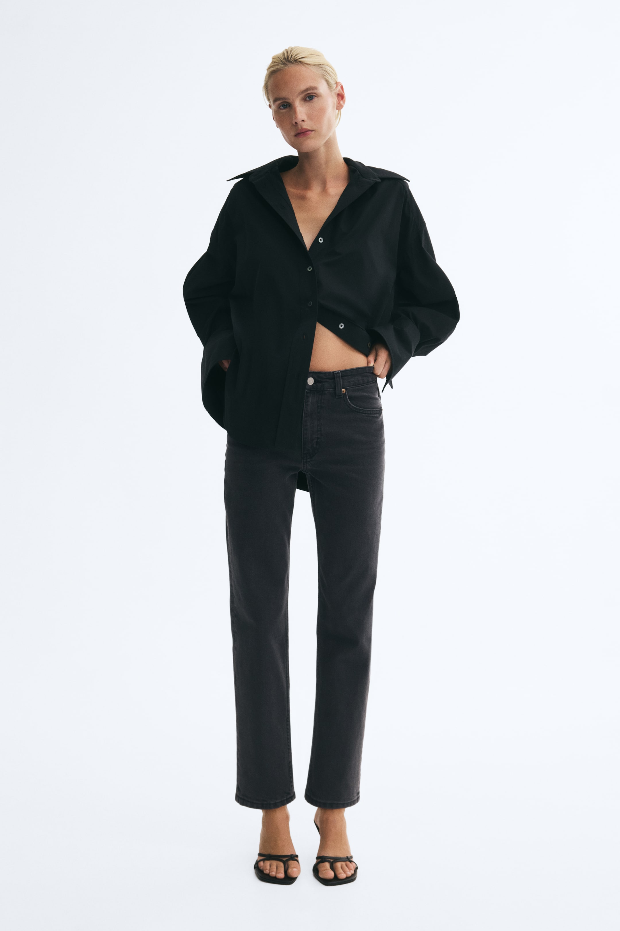 View larger image: Dark grey, high-waisted jeans with five pockets and a tapered leg are worn by the model, who is standing. The jeans are paired with an oversized black shirt, which the model is holding back to reveal the fitted waist and button closure.
