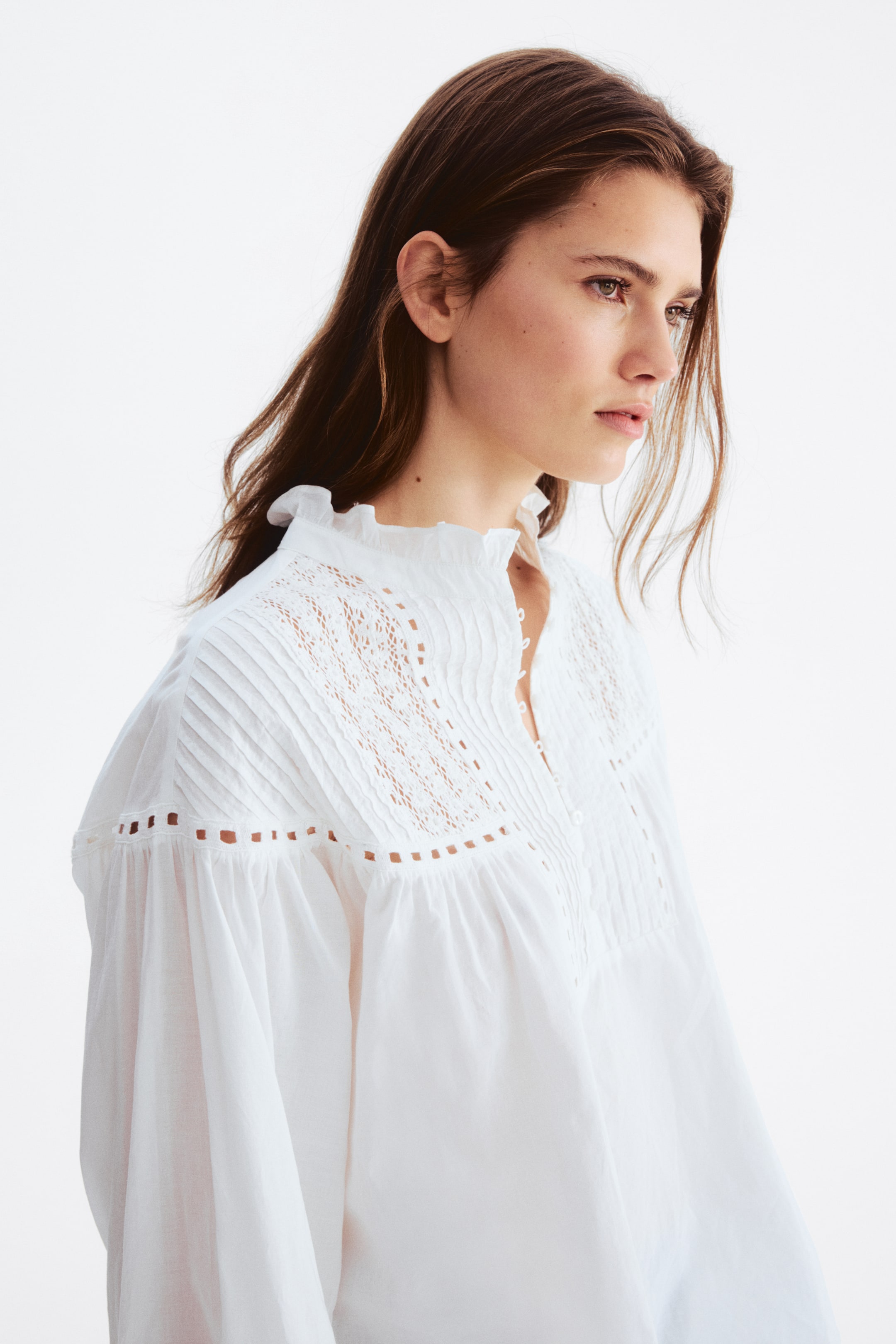 Agrandir l'image: Une blouse blanche est dotée d'un col montant à volants et d'une patte de boutonnage partielle. Le plastron avant est orné de plis nervurés verticaux, de panneaux en dentelle ajourée et d'une bordure horizontale à œillets, menant à des manches volumineuses froncées.