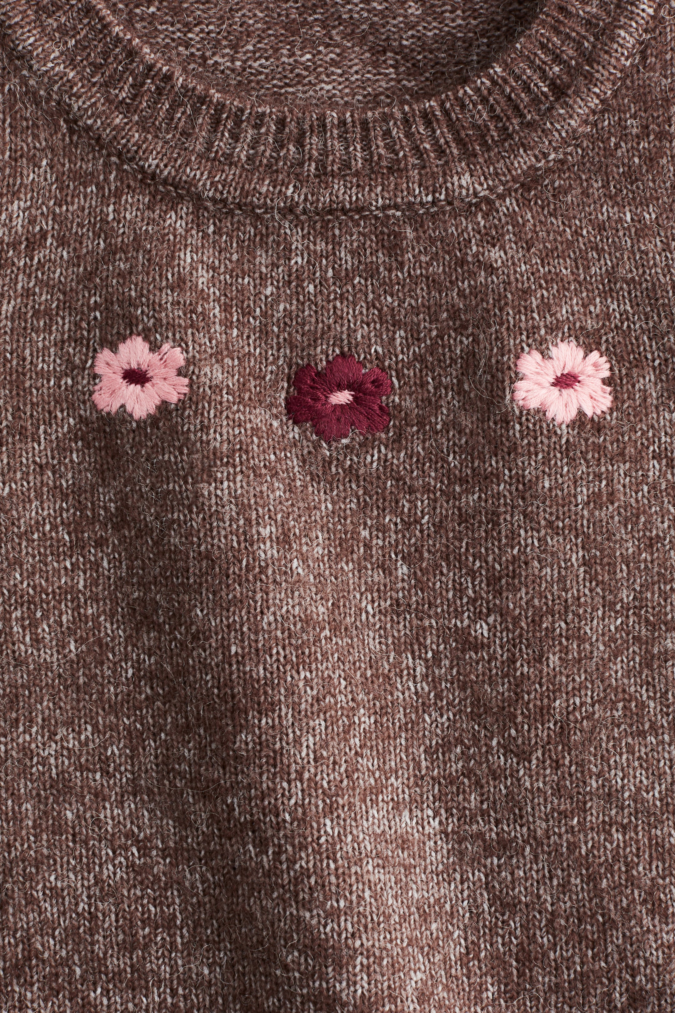 Ver imagem maior: Uma camisola de malha mesclada em castanho e branco apresenta uma gola redonda canelada e três flores bordadas, duas em rosa claro e uma em rosa escuro, dispostas horizontalmente na parte frontal do peito.
