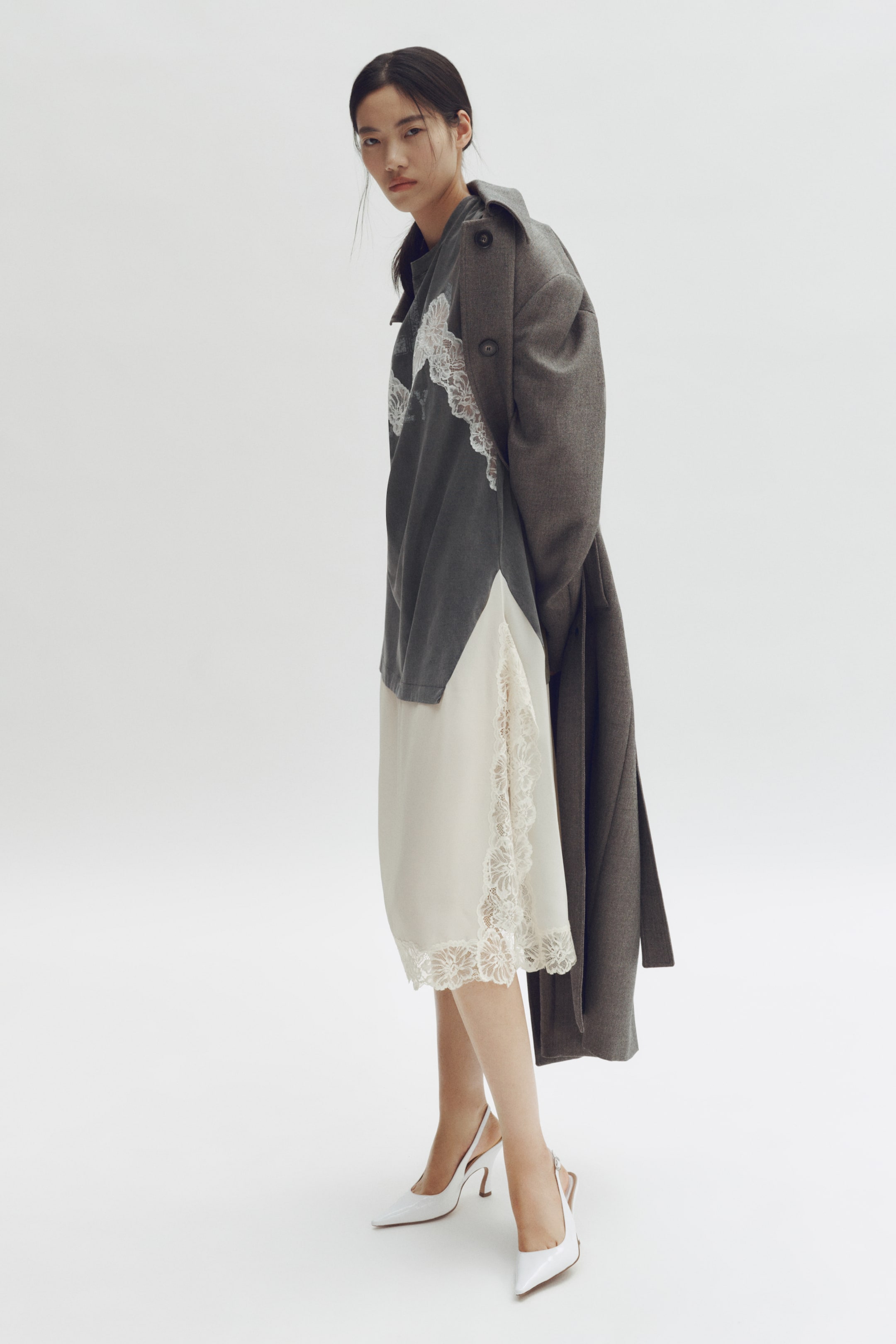 View larger image: A dark grey coat is draping over a grey long-sleeved top featuring white lace appliqué, which is layering over a cream slip dress with a lace-trimmed hem and side detail. White pointed-toe slingback heels are completing the outfit.
