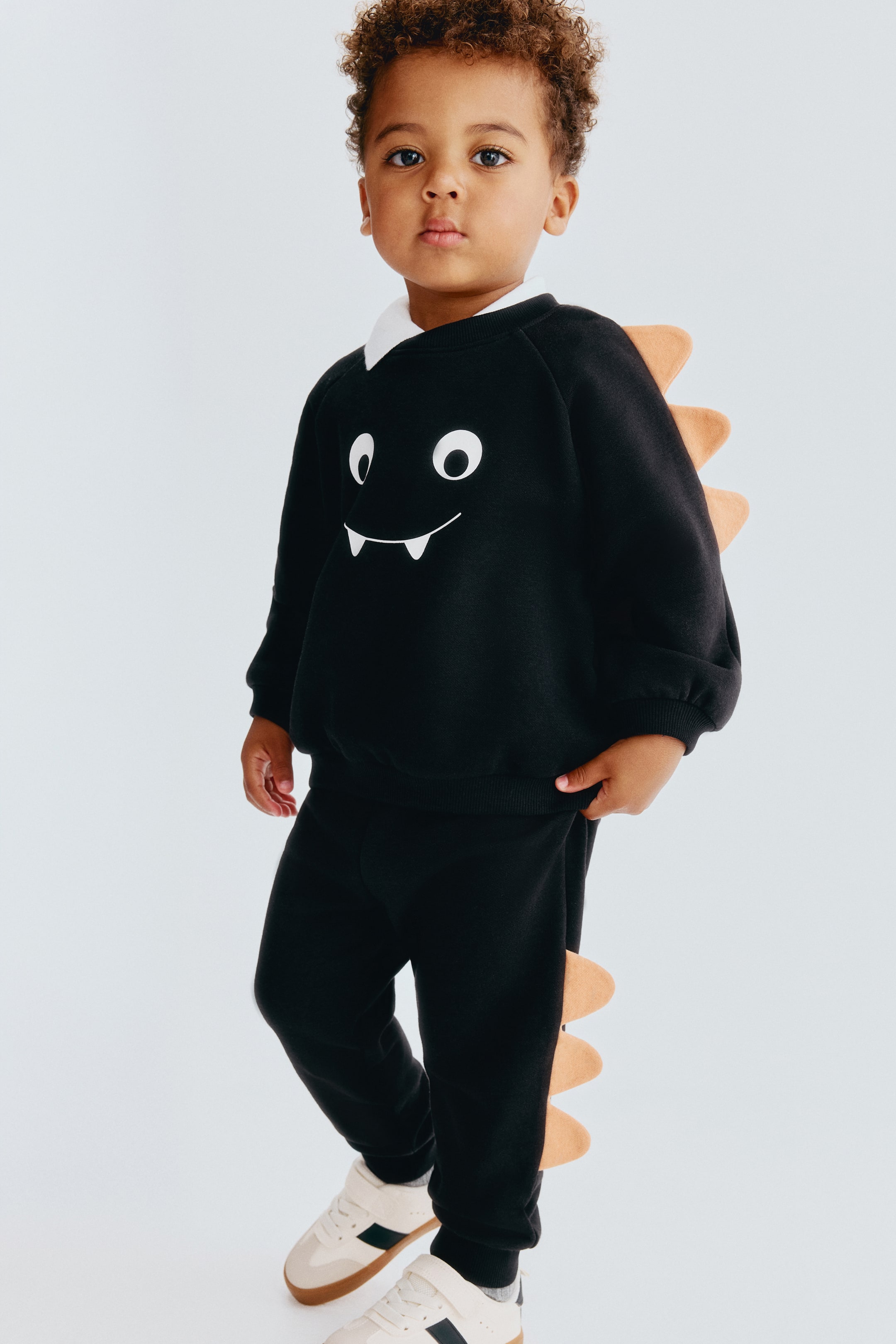 Größeres Bild anzeigen: Dieses von vorne gezeigte zweiteilige schwarze Set besteht aus einem langärmeligen Sweatshirt und einer passenden Jogginghose mit beigen, dinosaurierartigen Stoffspikes. Das Sweatshirt zeigt auf der Brust ein weißes Cartoon-Gesicht und ist über einem weißen Hemd mit Kragen getragen, während die Jogginghose entlang des unteren äußeren Beinbereichs mit passenden Spikes versehen ist.