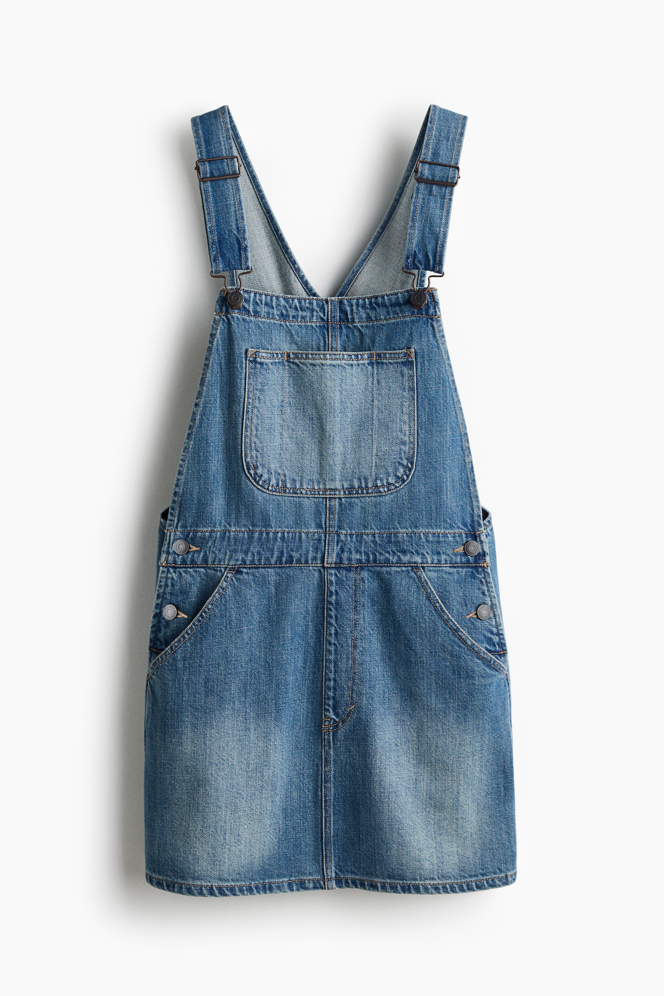 View larger image: A blue denim overall dress is showcased from the front, featuring adjustable shoulder straps with metal buckles, a large patch pocket on the bib, and two slanted side pockets with buttons at the waist.