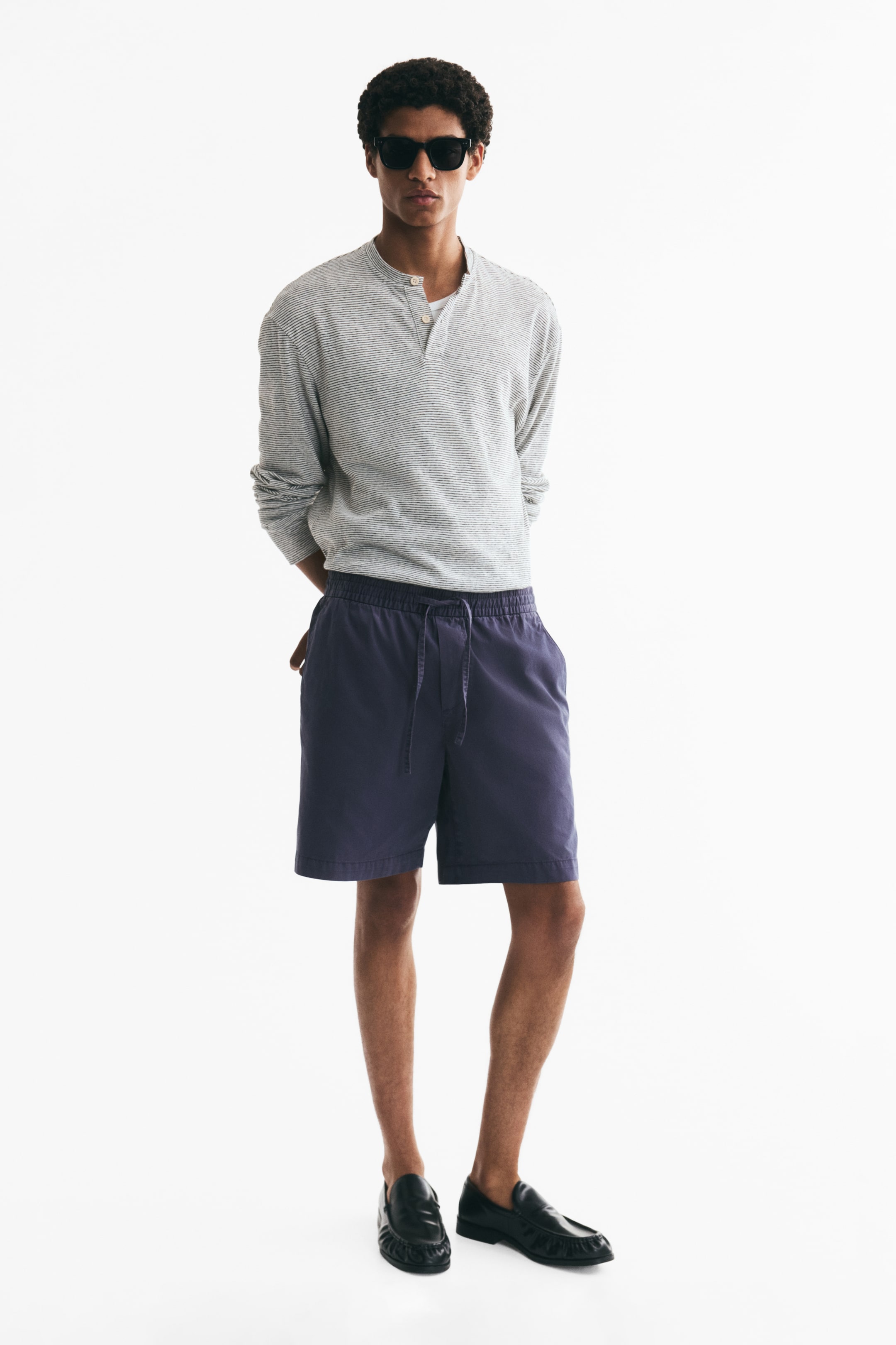 Xem ảnh lớn hơn: Người mẫu đang mặc quần short vải cotton twill màu xanh navy. Thiết kế có cạp chun rút dây, túi chéo hai bên và một túi viền sau, mang phom dáng thoải mái khi người mẫu đứng với một tay đặt trong túi bên.