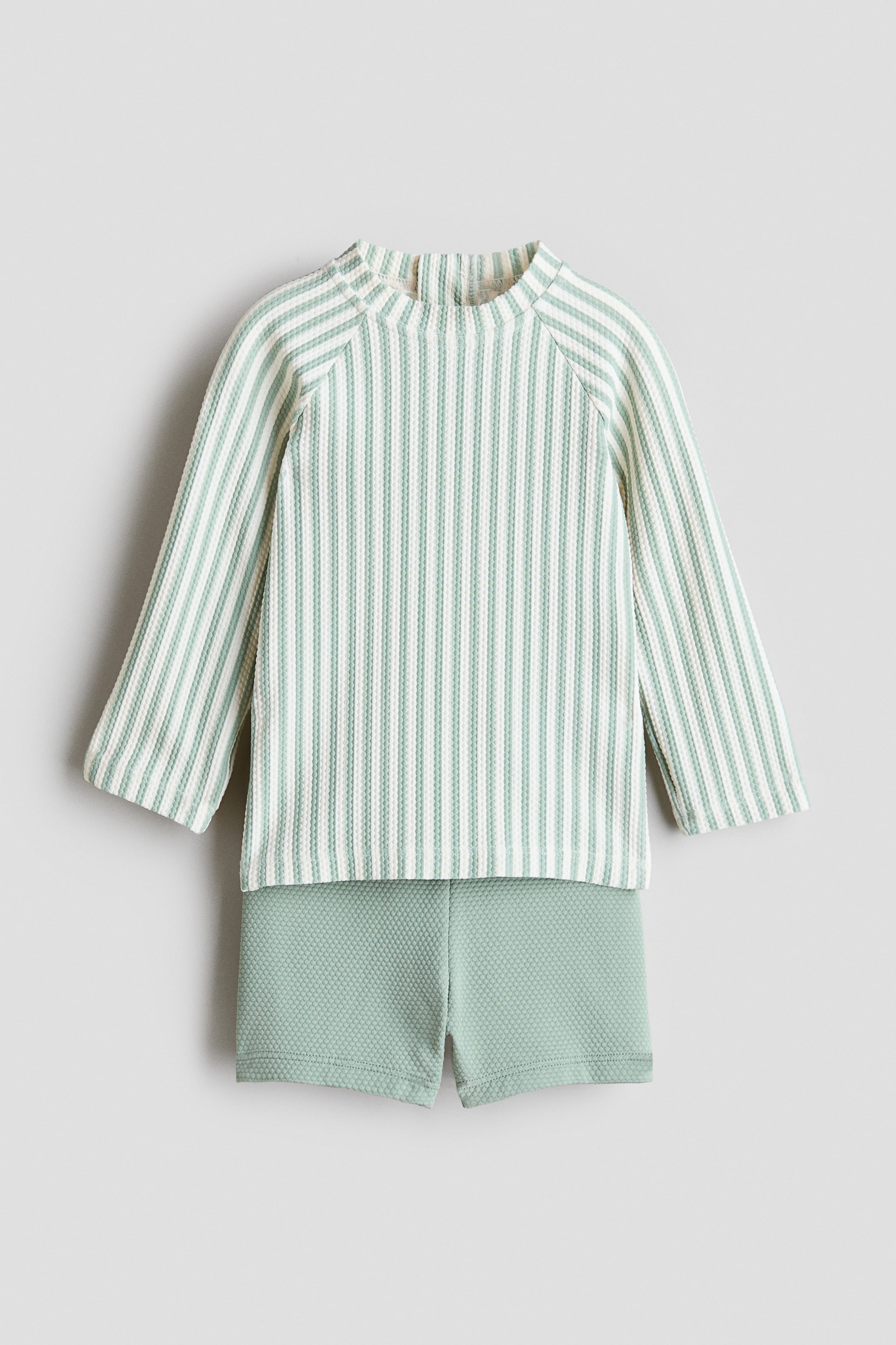 Agrandir l'image: Ensemble deux pièces présentant un haut à manches longues raglan, à encolure ronde haute et à rayures verticales vert pâle et blanc cassé, associé à un short uni vert pâle. Les deux pièces sont en maille gaufrée texturée.