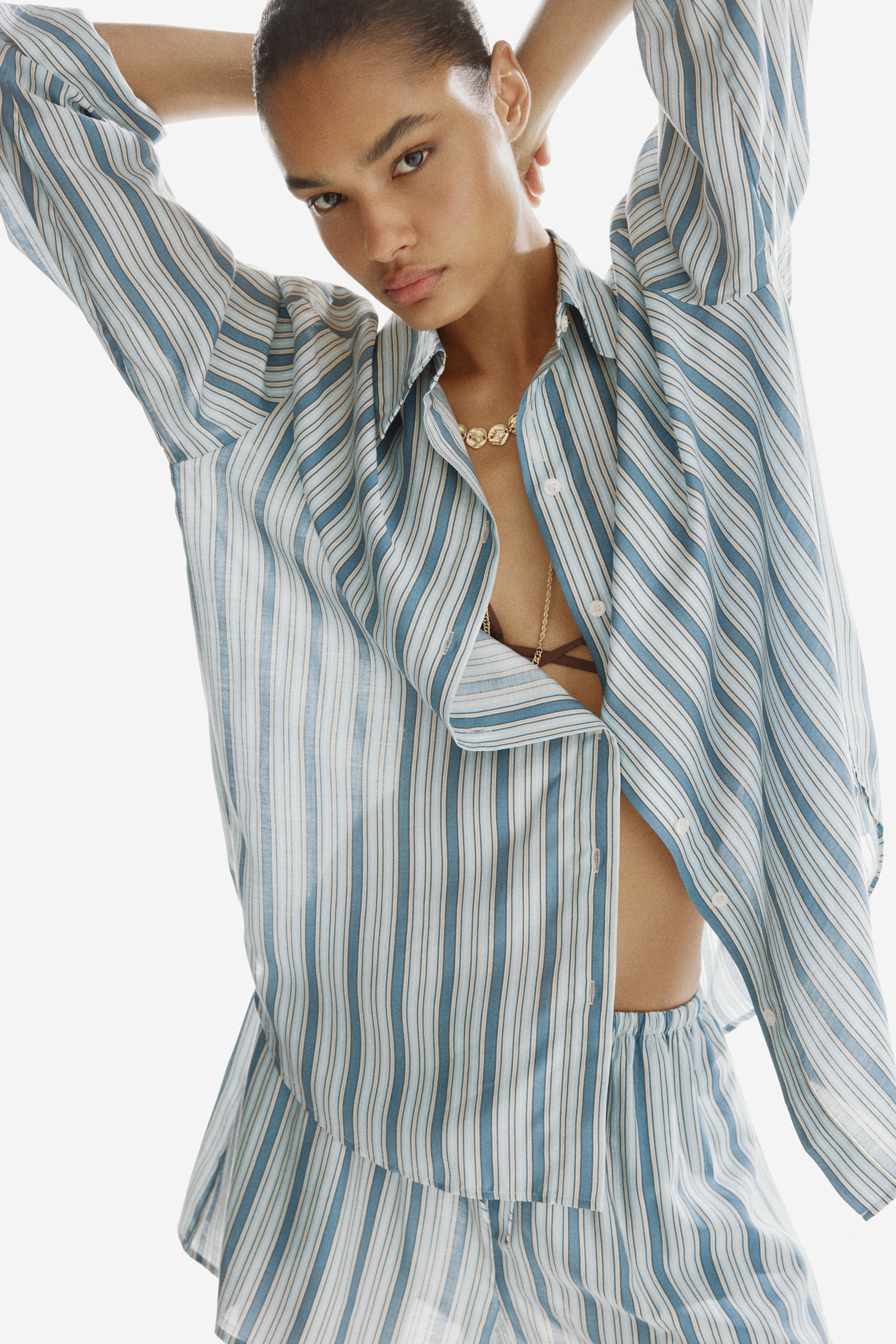 Ampliar la imagen: La modelo lleva puesta una camisa a rayas de gran tamaño con un ligero brillo, que cuenta con un cuello, hombros caídos, mangas largas con puños abotonados y un dobladillo redondeado. La modelo levanta los brazos, lo que hace que la parte delantera parcialmente desabotonada de la camisa se abra, revelando un sostén y un collar, y también lleva puestos shorts a rayas a juego.
