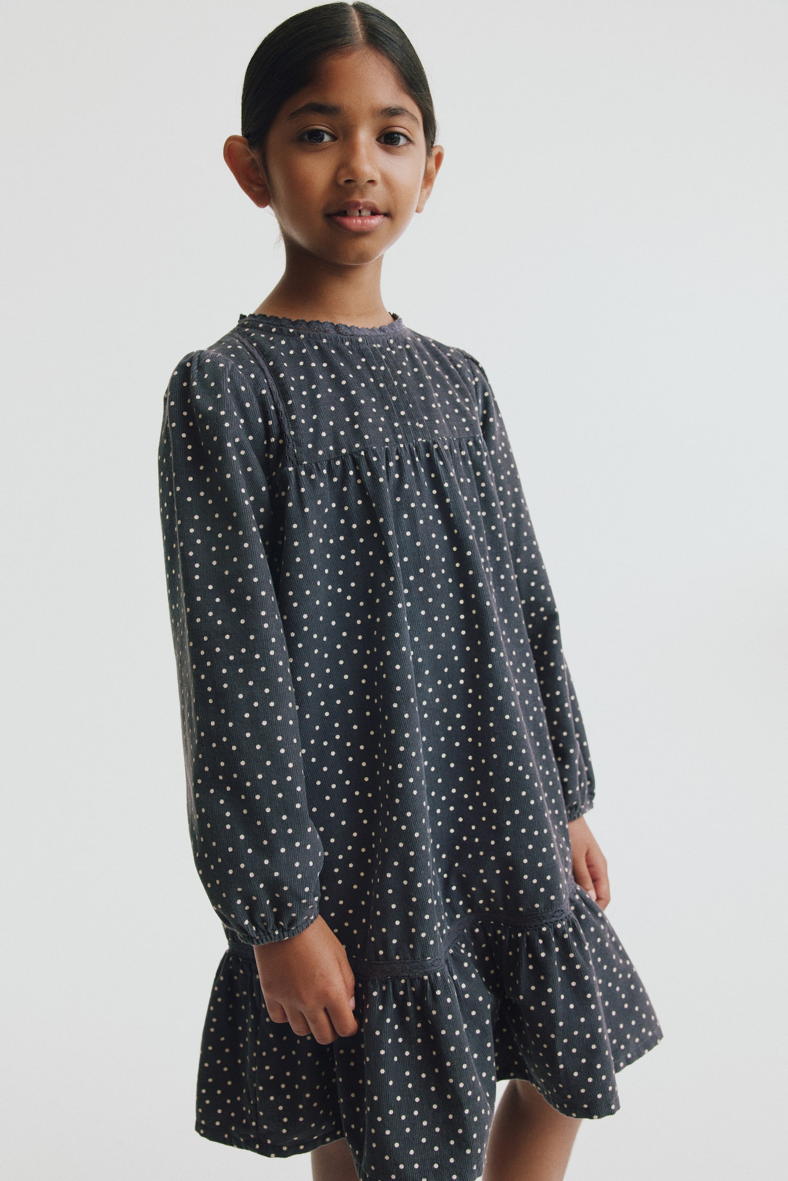 A dark grey A-line corduroy dress featuring a white polka-dot print is shown from the front. The dress is designed with a round, lace-trimmed neckline, a front yoke with gathers, long balloon sleeves with narrow elasticised cuffs, and a gathered ruffle at the hem. - 1