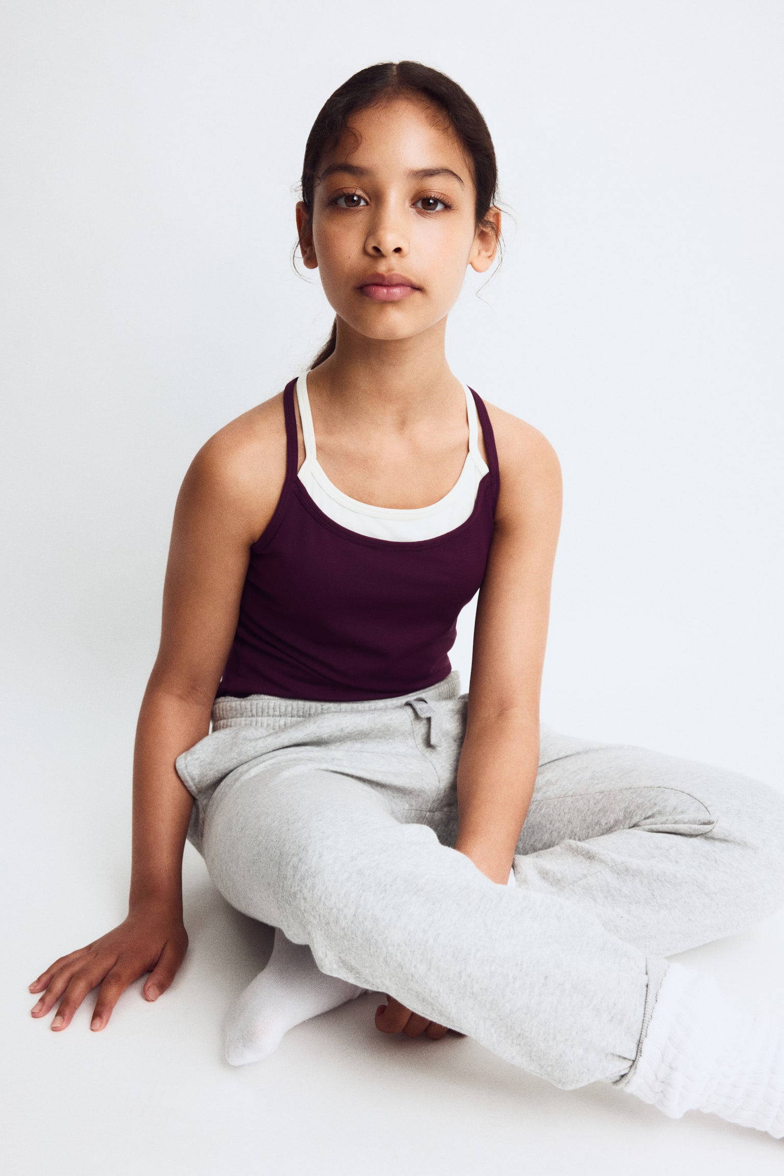 A plum-coloured tank top is showcasing a layered design from the front. It features thin straps and a scoop neckline, with an off-white inner layer revealing criss-cross straps at the back. - 1