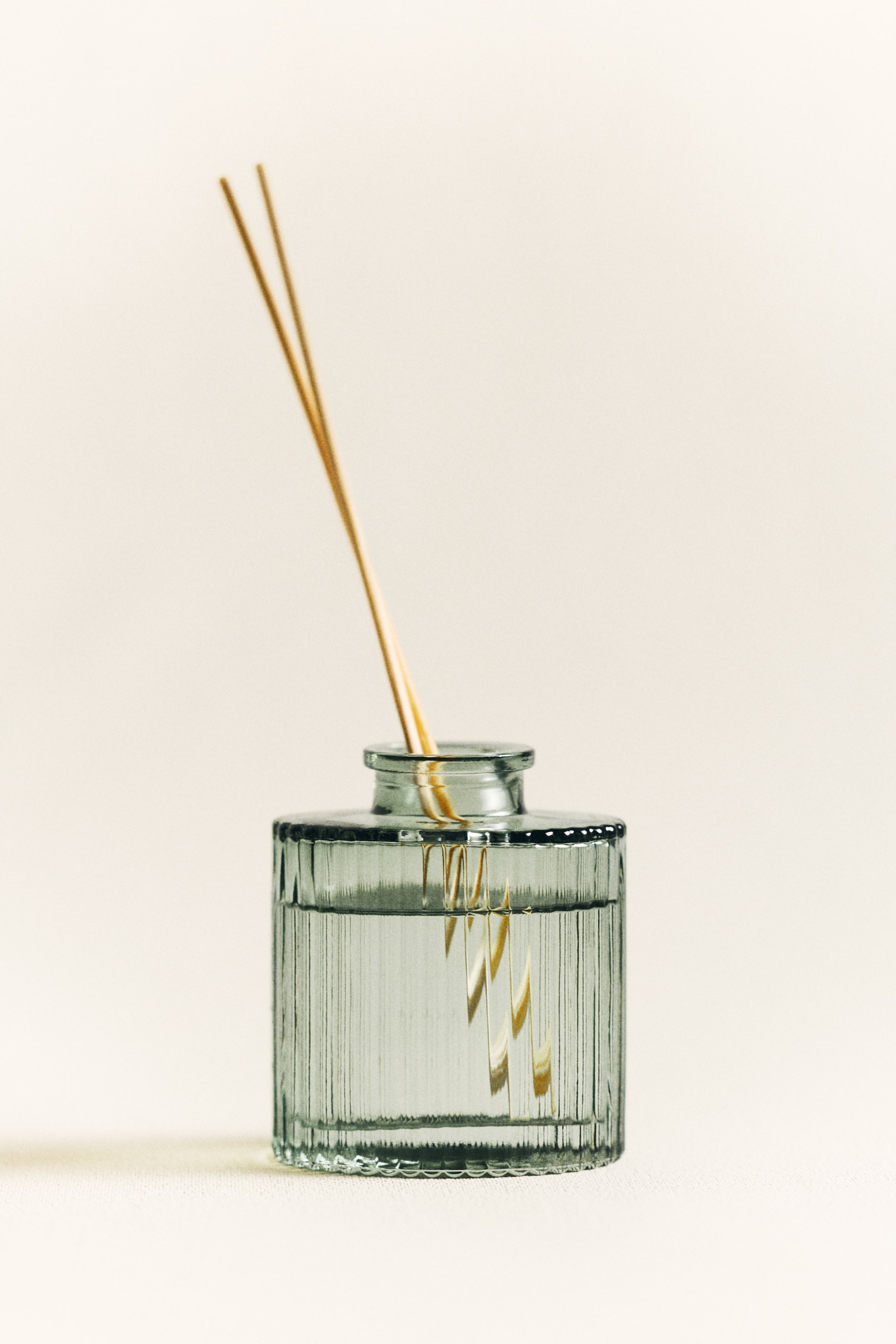 View larger image: A reed diffuser features a rectangular, fluted glass bottle filled with a clear liquid. Several natural-colored reeds stand upright and partially submerged in the liquid, fanning slightly outward.