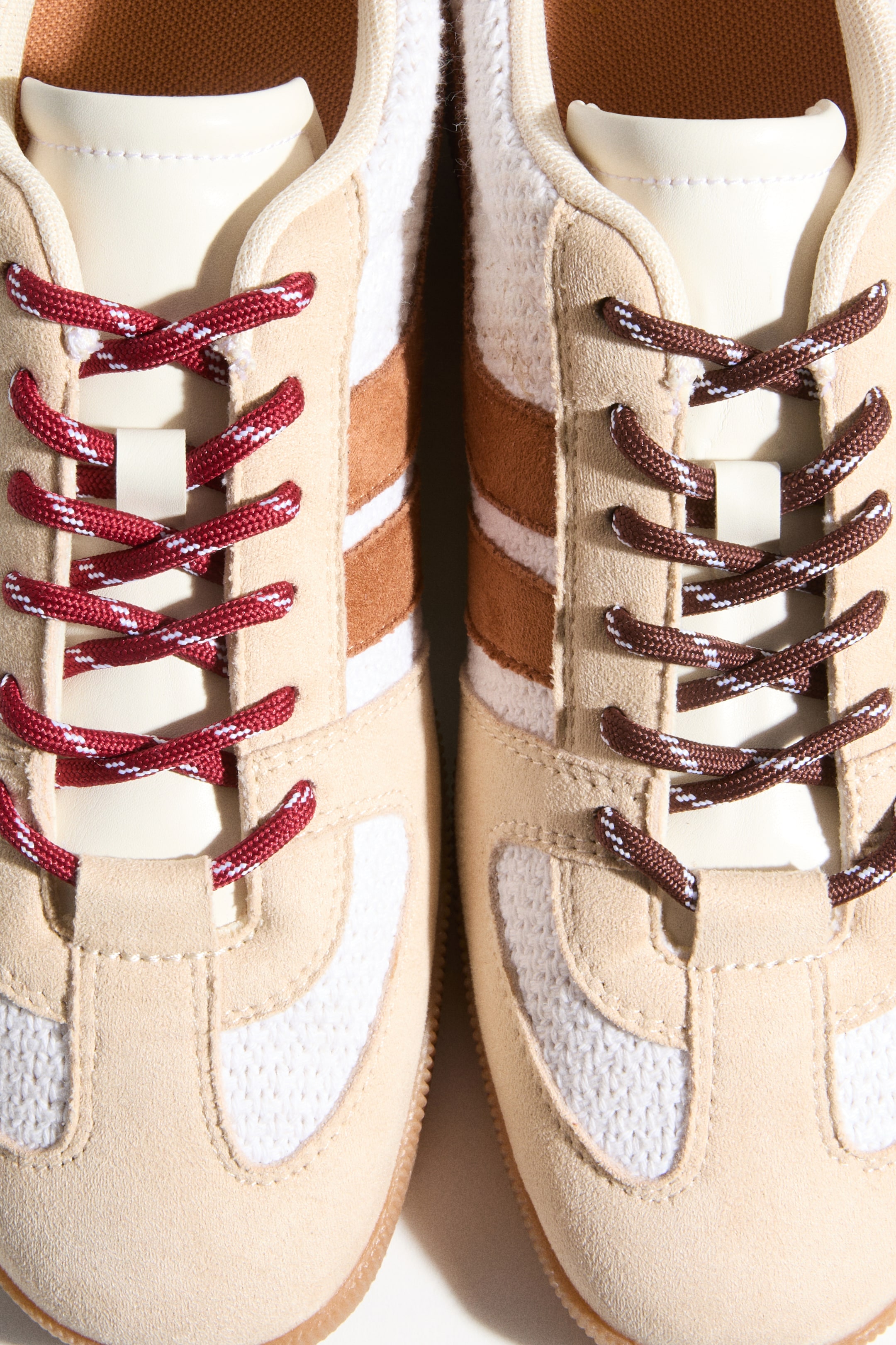 Voir une image plus grande: Une paire de baskets est présentée en vue de dessus, avec des dessus en similicuir beige et des inserts en maille blanche sur le bout et les panneaux latéraux. Des rayures horizontales marron et blanches ornent le milieu du pied, et chaque chaussure est lacée avec des lacets rayés contrastants : rouges et blancs à gauche, et marron et blancs à droite, tous deux croisés sur les languettes visibles de couleur crème.