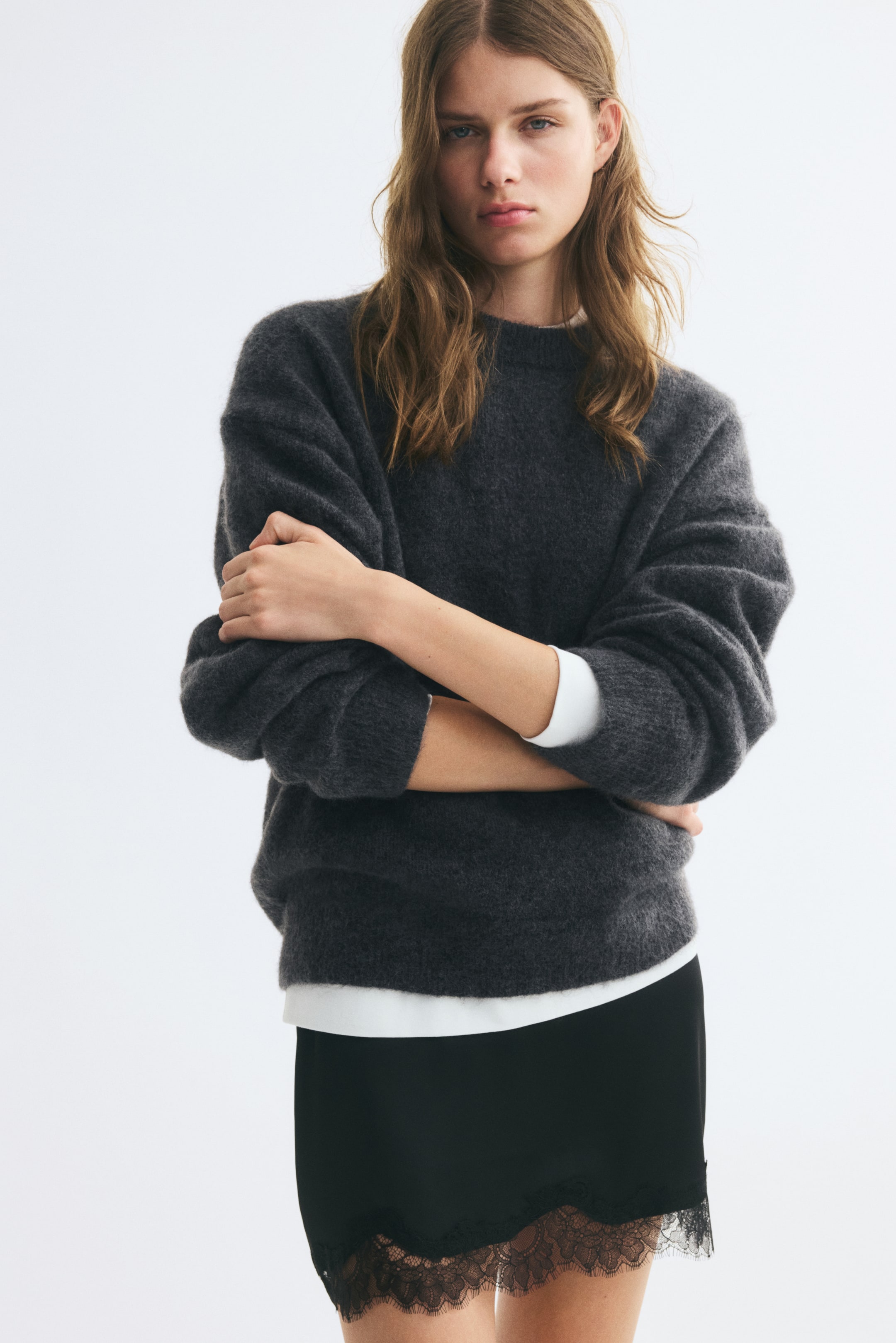 View larger image: A dark grey, soft-knit jumper featuring a round ribbed neckline, dropped shoulders, and ribbed cuffs and hem is being worn by the model. The jumper is layered over a white long-sleeve top and paired with a black lace-trimmed skirt, with the model crossing their arms.