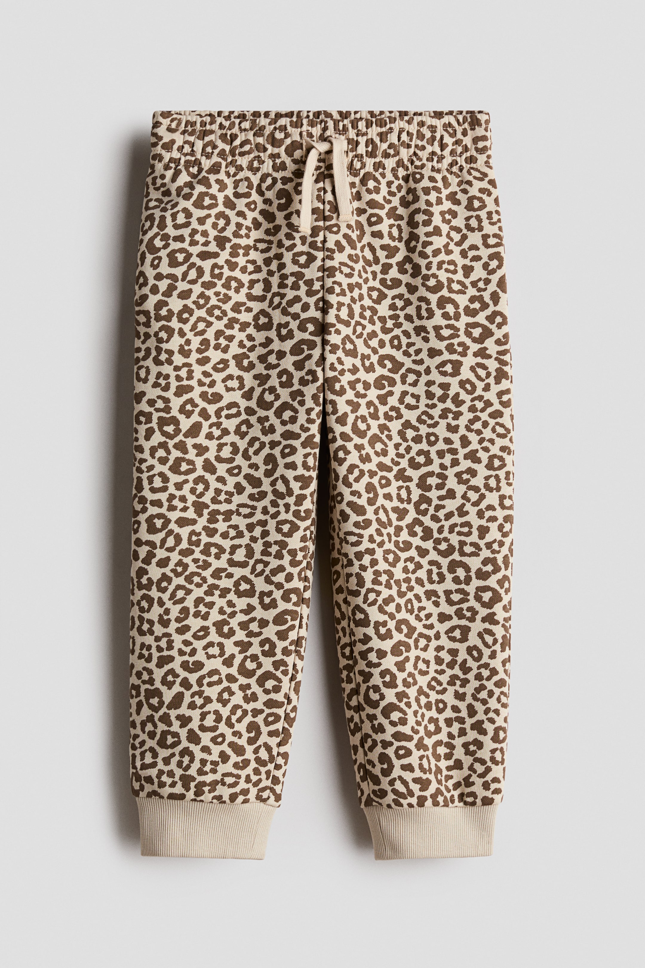 View larger image: A pair of track pants featuring an all-over brown leopard print is shown from the front, with an elastic waistband, a cream-colored drawstring, and solid cream-colored ribbed cuffs at the ankles.