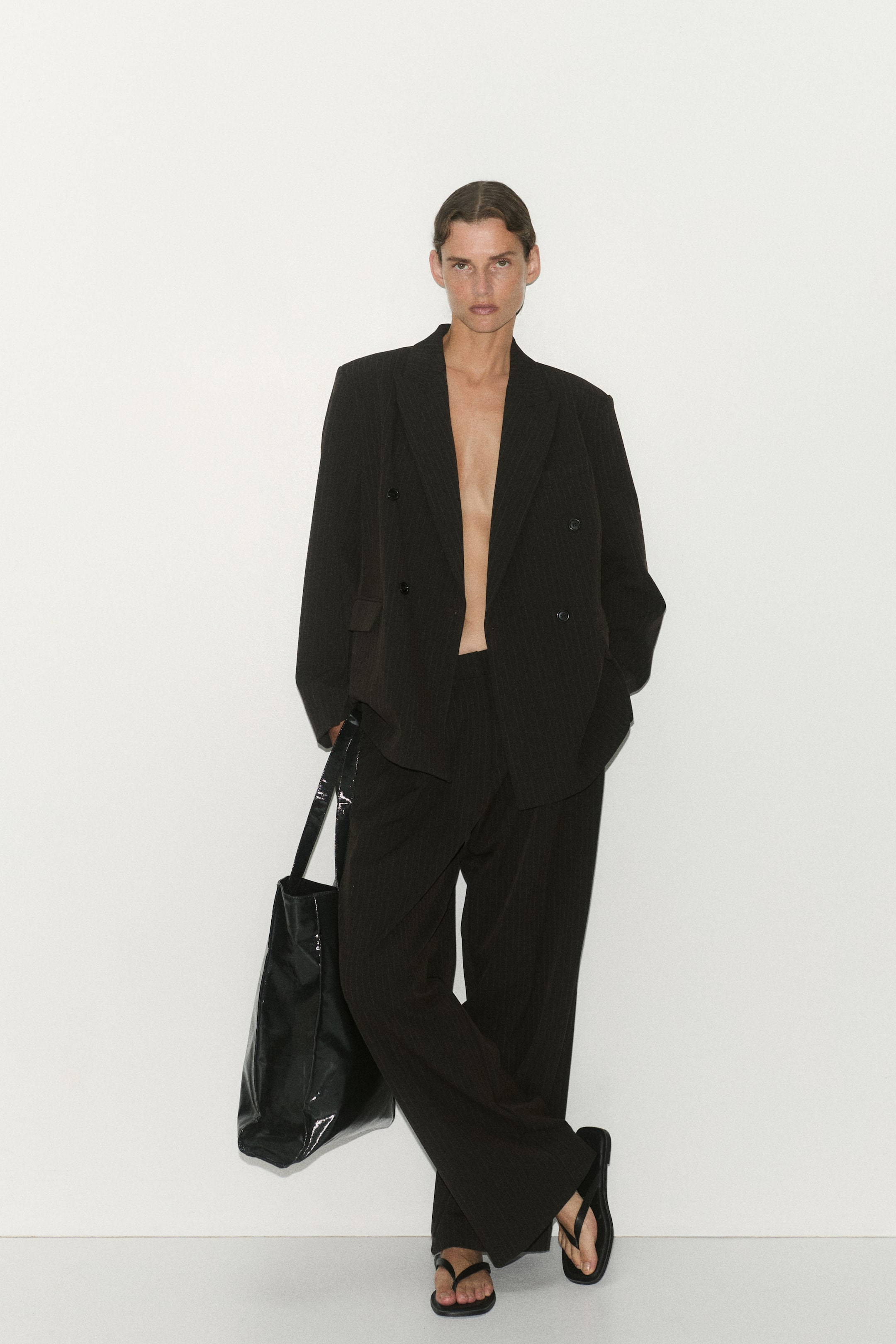 Visualizza immagine più grande: Pantaloni dal taglio morbido e sartoriale in tessuto a righe sottili scuro, con silhouette ampia a gamba larga, mostrati frontalmente con pinces anteriori e pieghe lungo le gambe. I pantaloni a vita alta sono abbinati a un blazer doppiopetto coordinato, lasciato sbottonato, per valorizzarne la struttura rilassata ma raffinata.