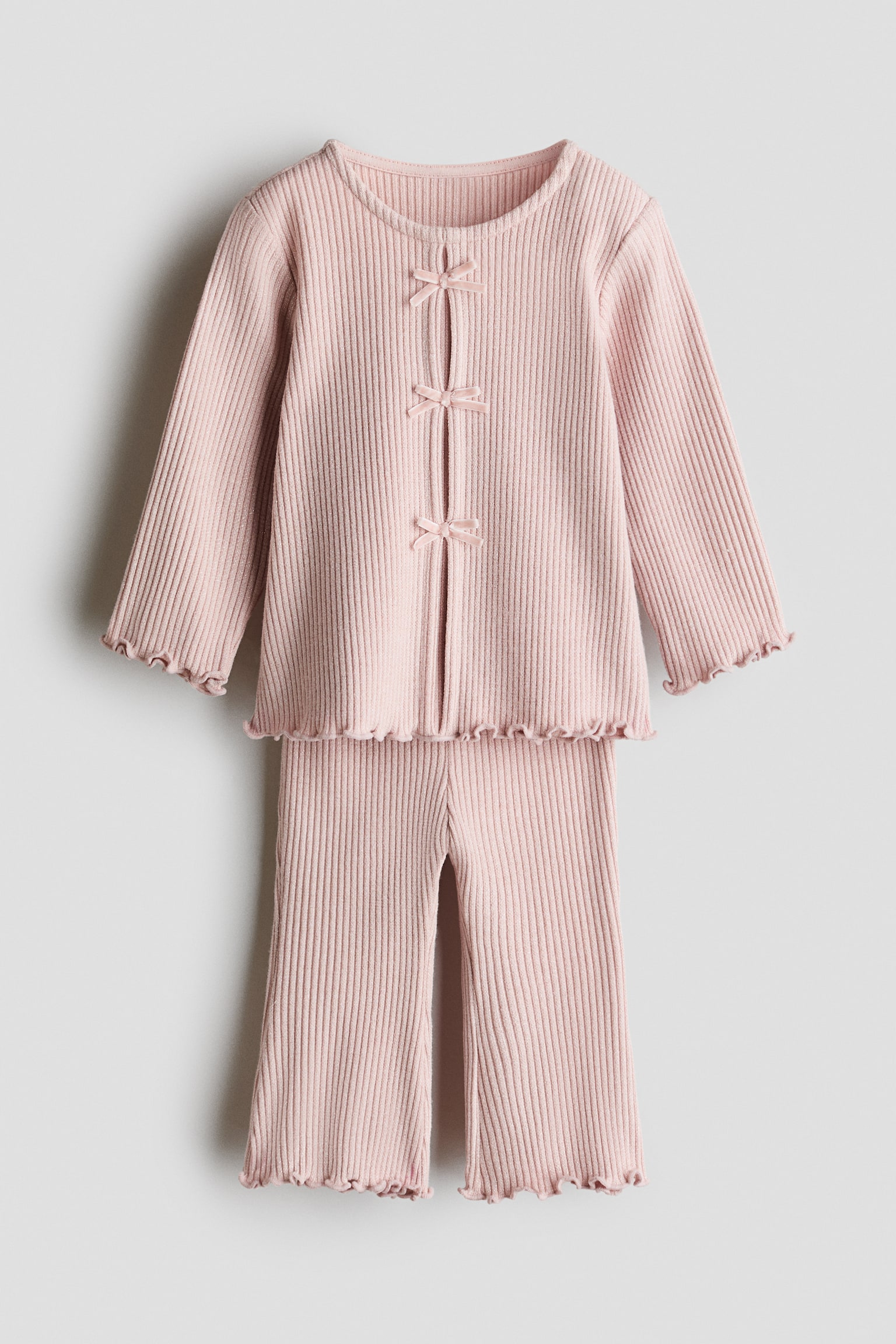 Un ensemble deux piÃ¨ces en tricot cÃ´telÃ© rose pÃ¢le est prÃ©sentÃ© de face, composÃ© dâun haut Ã  manches longues et dâun pantalon assorti. Le haut prÃ©sente trois dÃ©licats nÅuds en ruban le long de la fermeture Ã  lâavant ainsi que des finitions Ã  bord ondulÃ© aux poignets et Ã  la taille, tandis que le pantalon affiche une jambe Ã©vasÃ©e avec des ourlets volantÃ©s assortis.