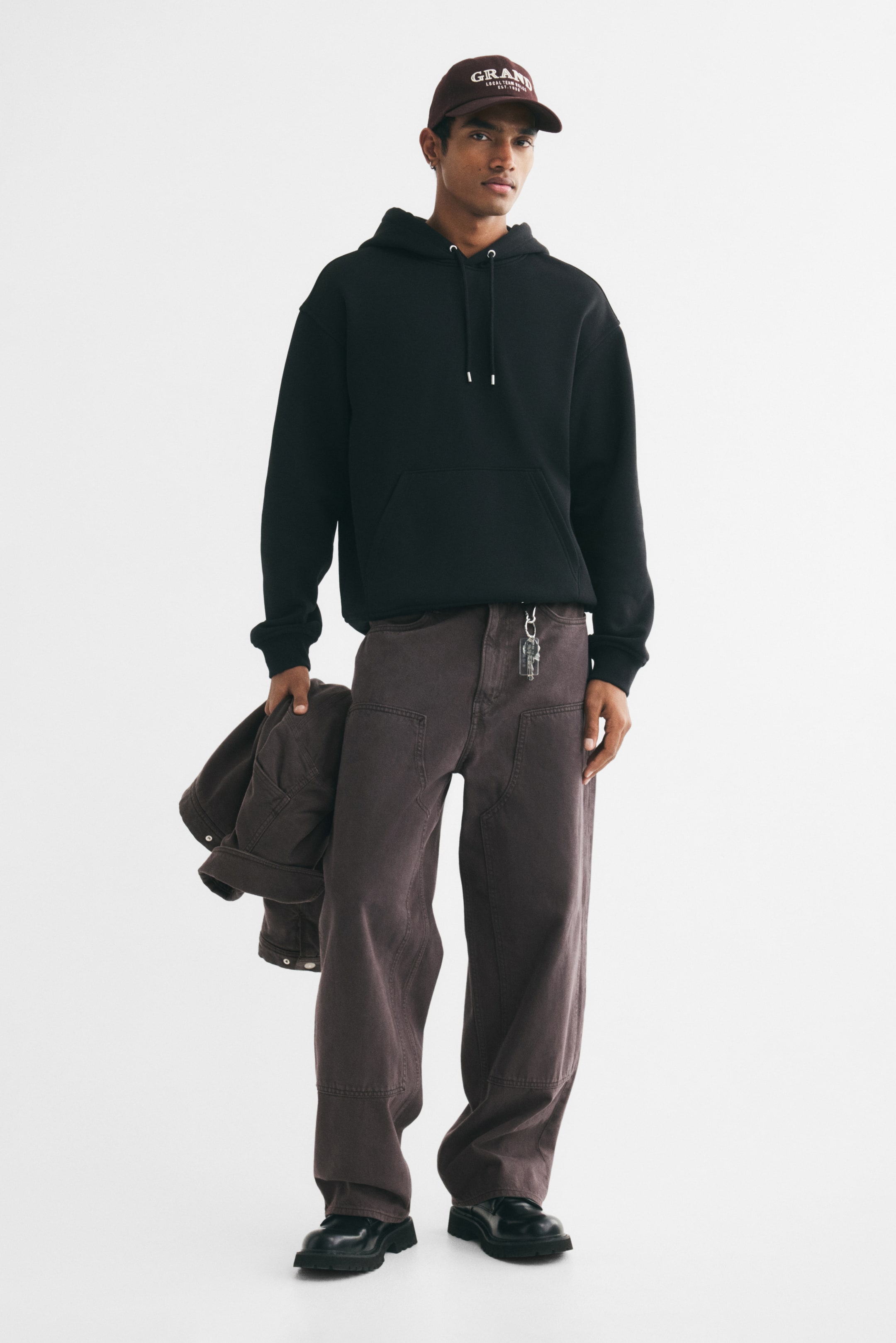 View larger image: A black hoodie with a drawstring and kangaroo pocket is being worn by the model, paired with wide-leg brown utility pants. The model is holding a matching brown jacket in one hand while wearing a dark brown baseball cap and black shoes.
