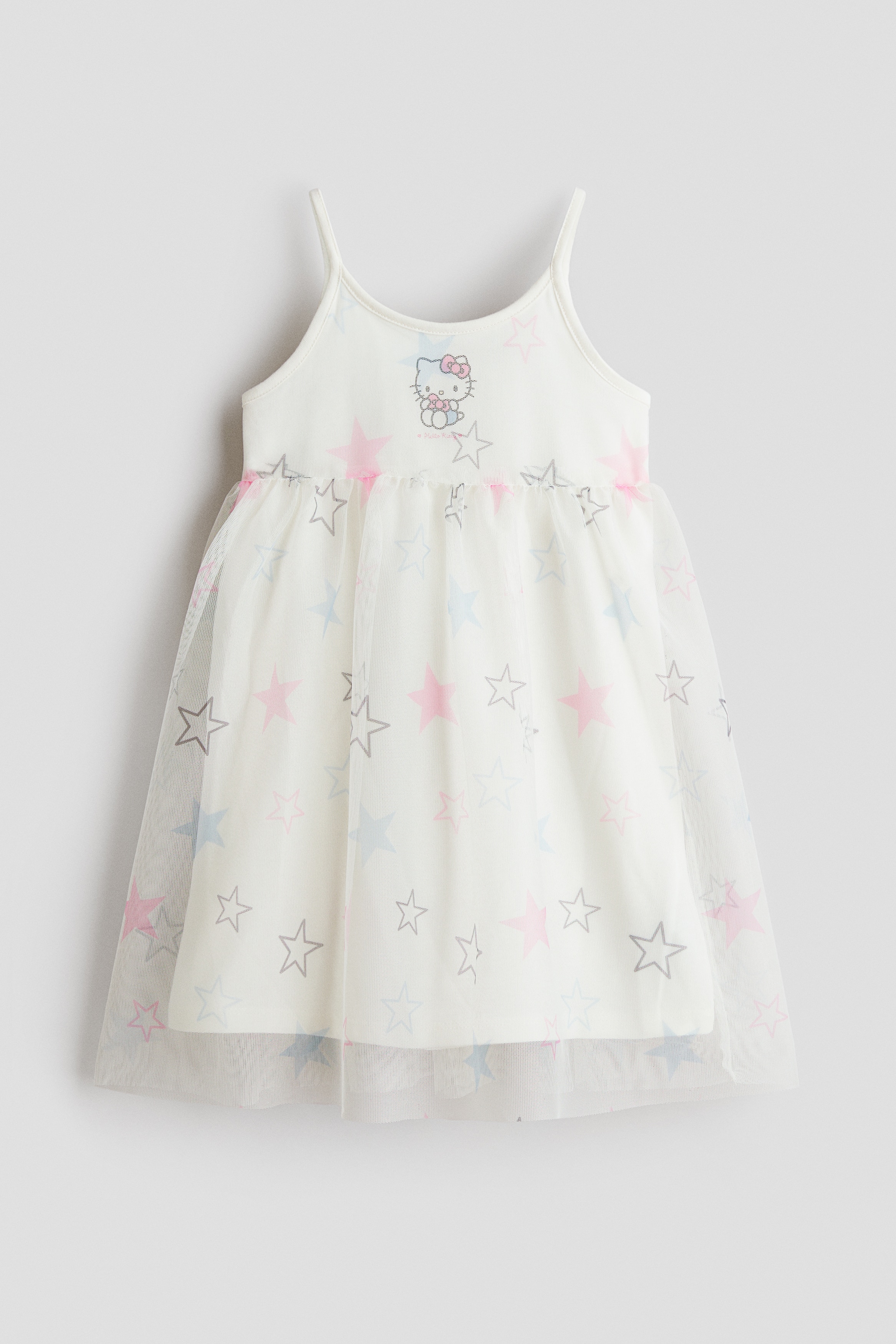 A white jersey dress with narrow shoulder straps and a round neckline is worn by the model. The dress features a printed cartoon character on the chest and a flared skirt with a sheer tulle overlay decorated with a multicoloured star print.