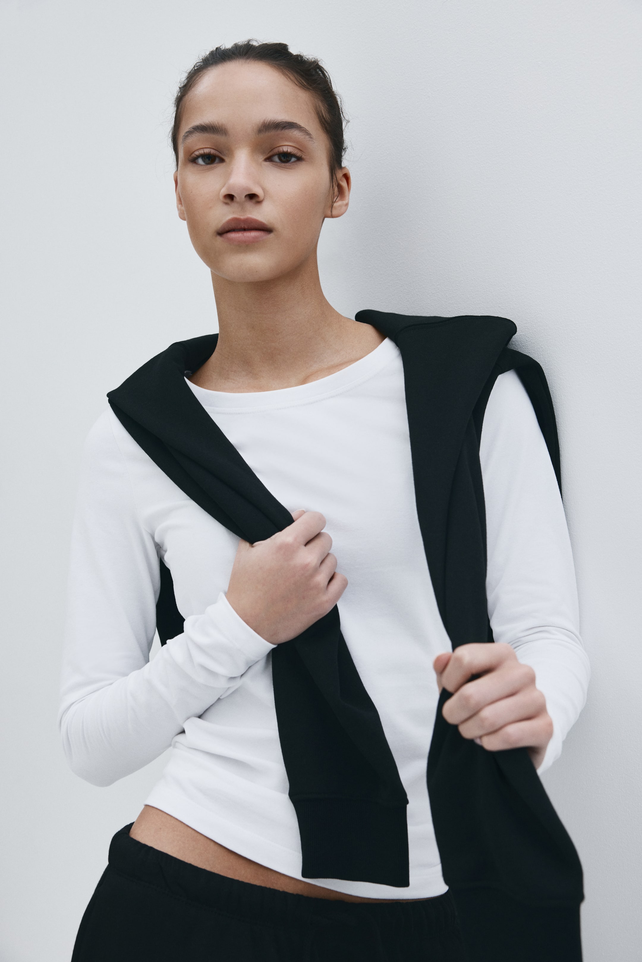 View larger image: A black garment is draped over the model's shoulders, and the model is holding its ends with both hands.
