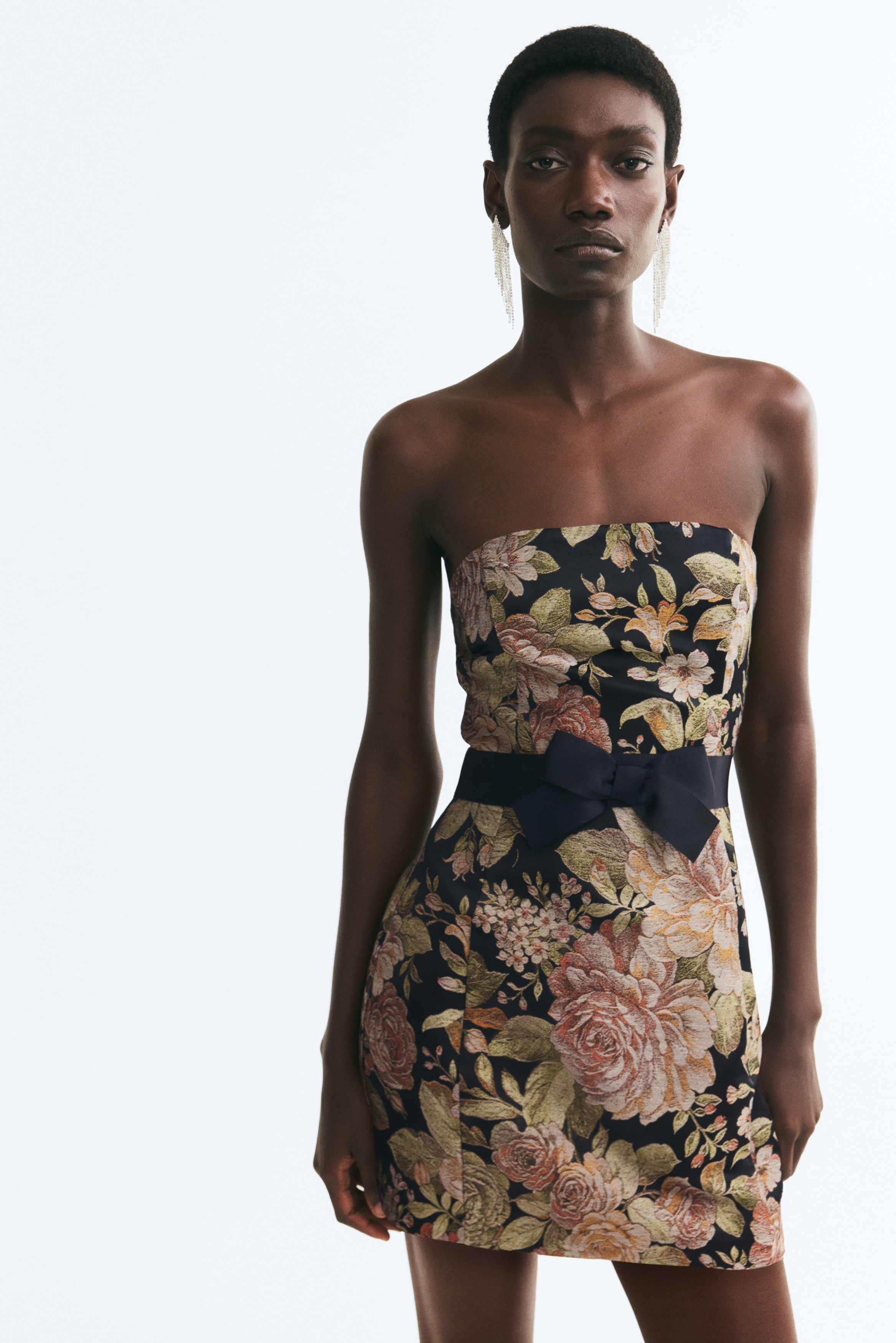 A strapless mini dress is shown from the front, featuring a metallic floral jacquard pattern in warm gold, bronze and muted green tones on a black background. The structured design is cinched at the waist with a black satin ribbon belt tied into a central bow.