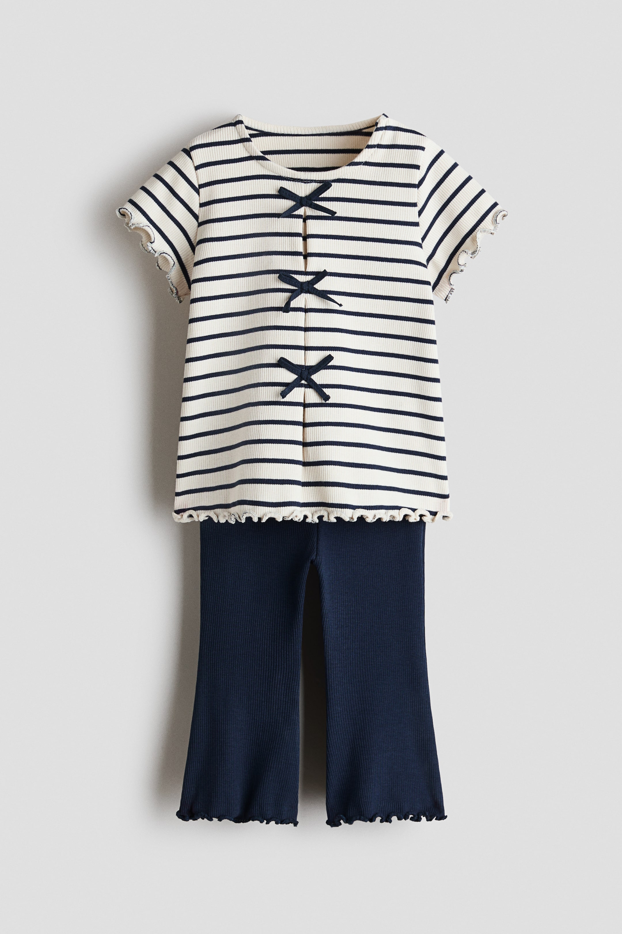 View larger image: A two-piece ribbed outfit showcases a cream and navy striped top paired with solid navy pants, presented front-facing. The short-sleeved top features a round neckline, ruffled sleeve edges, and a ruffled bottom hem, accented by three navy bows adorning its center front. The navy pants display a wide-leg style with ruffled hems.