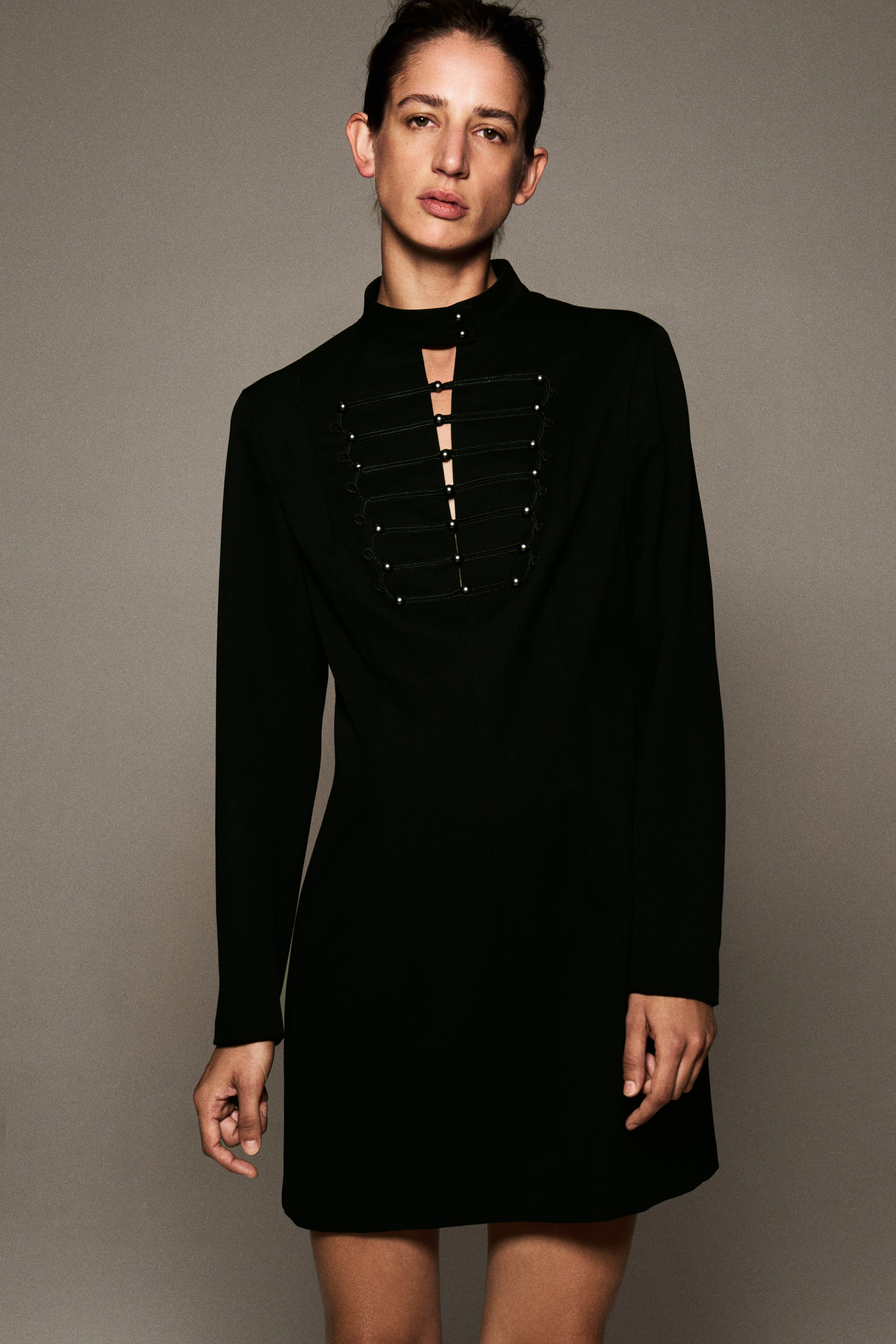 Ampliar la imagen: La modelo lleva un vestido corto negro entallado, con cuello alto, broche abotonado en el cuello y una abertura al frente. El vestido de manga larga presenta en el pecho detalles de cordones horizontales decorativos y pequeños botones metálicos.