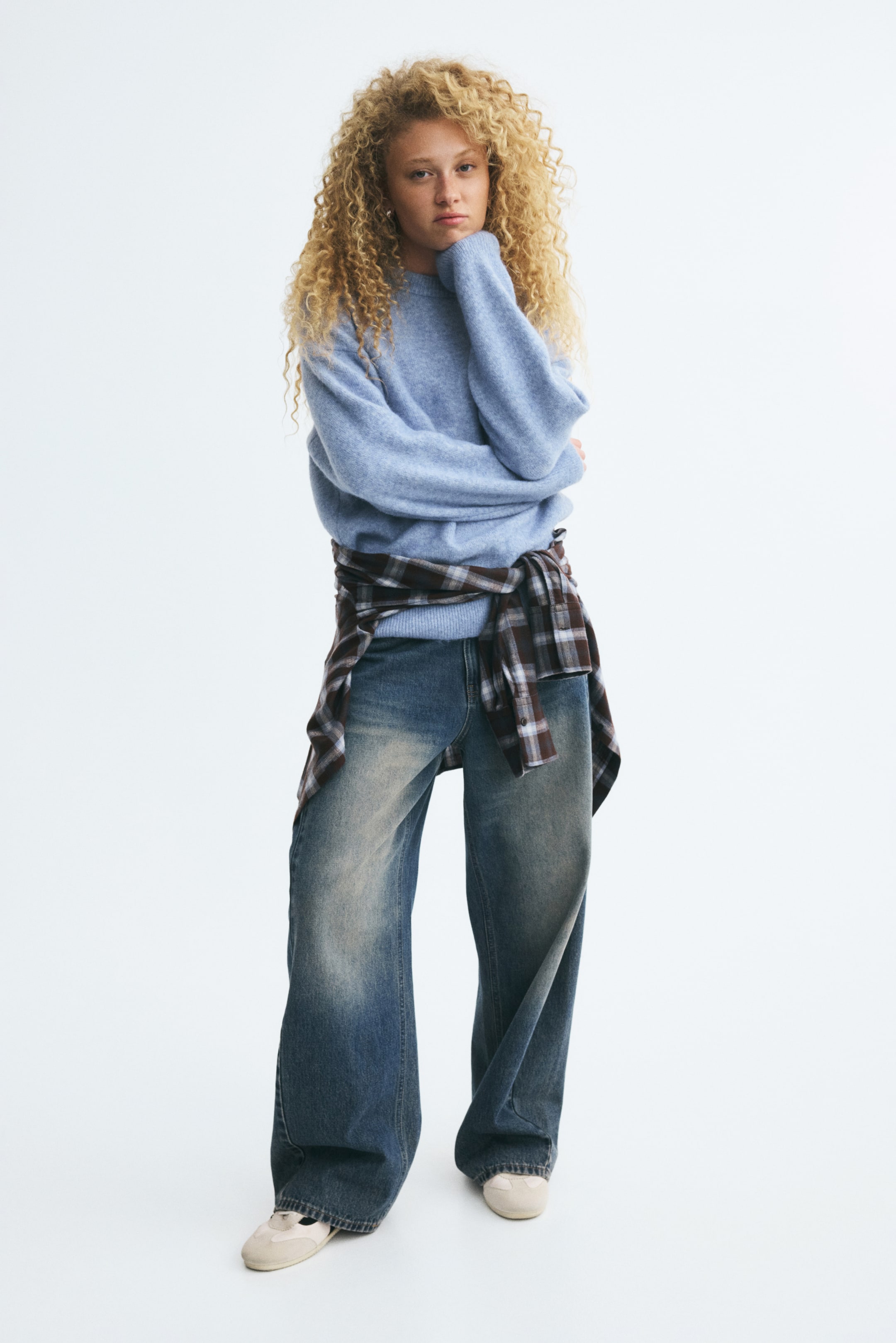 Visualizza immagine più grande: Un maglione in maglia azzurro chiaro dalla vestibilità morbida, con scollo rotondo e spalle scese, indossato dalla modella, che appoggia il mento sulla mano coperta dalla manica. Il maglione è abbinato a jeans a gamba larga e a una camicia a quadri annodata in vita.