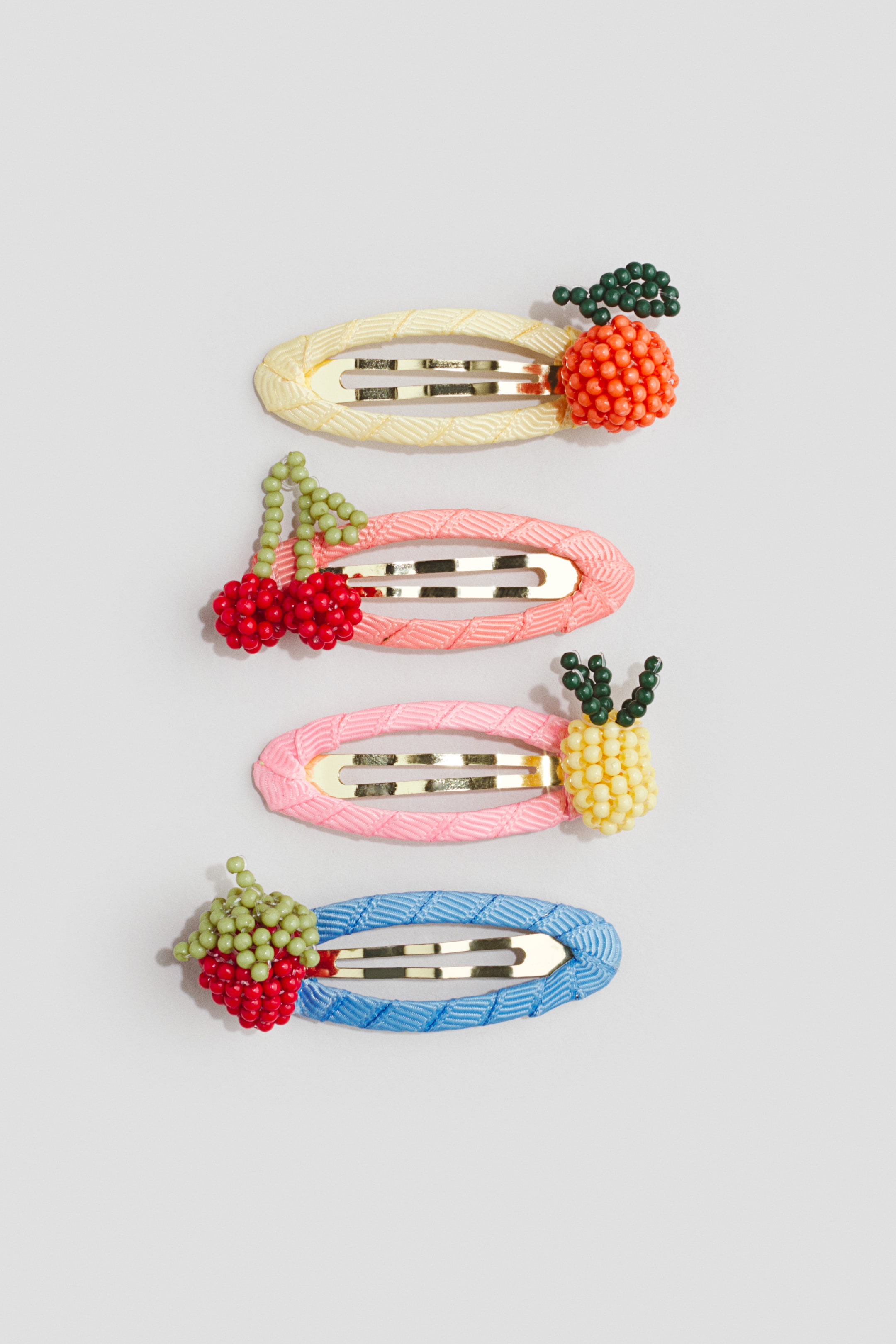 View larger image: Four oval snap hair clips are arranged vertically, each featuring a textured fabric-wrapped base and a unique beaded fruit decoration. A yellow clip is topped with an orange, a pink clip with cherries, a light pink clip with a pineapple, and a blue clip with a strawberry.
