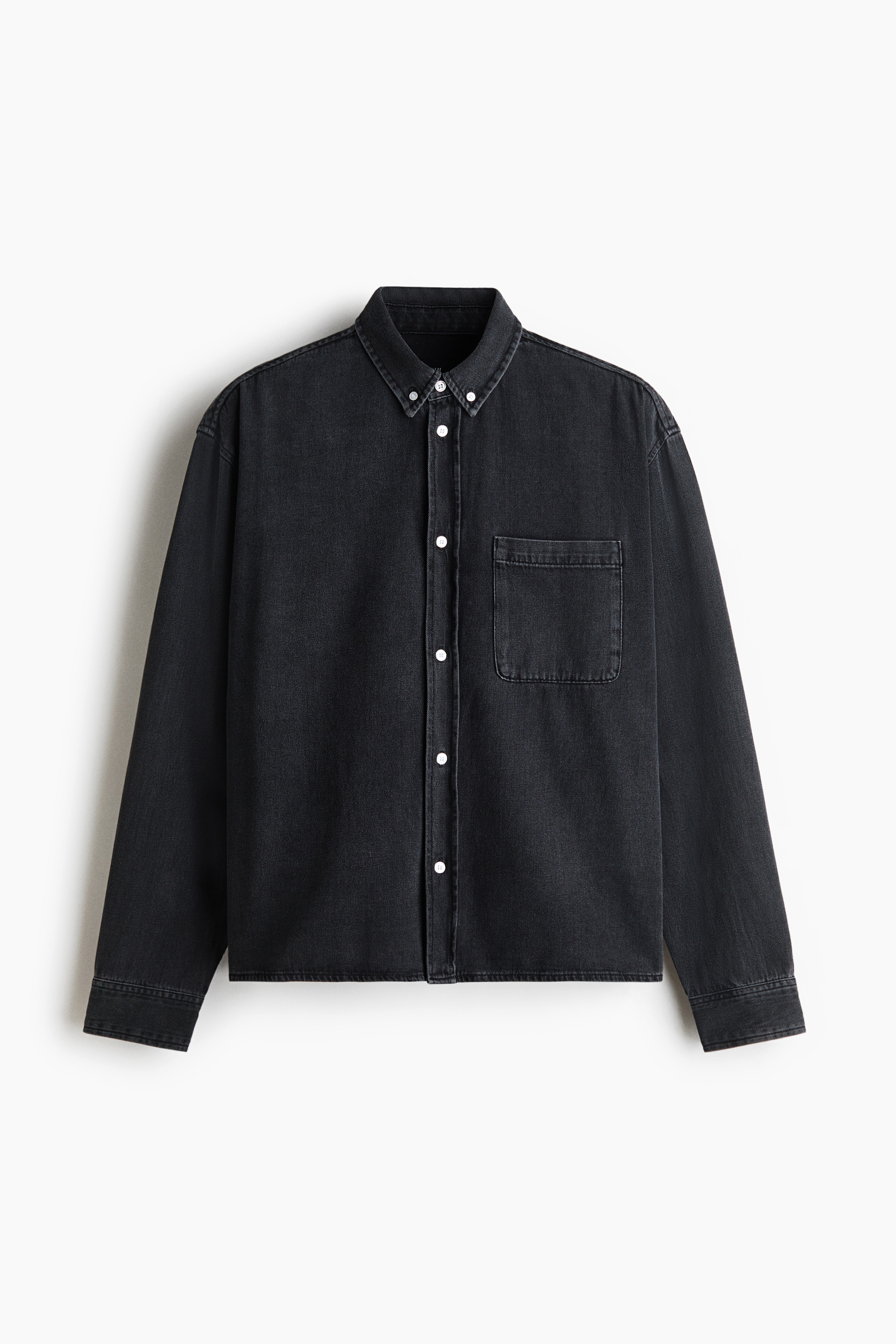 A dark grey cotton denim shirt is being worn open by the model over a light grey polo shirt and black trousers. The relaxed-fit shirt features a button-down collar, white buttons, and a single patch chest pocket.