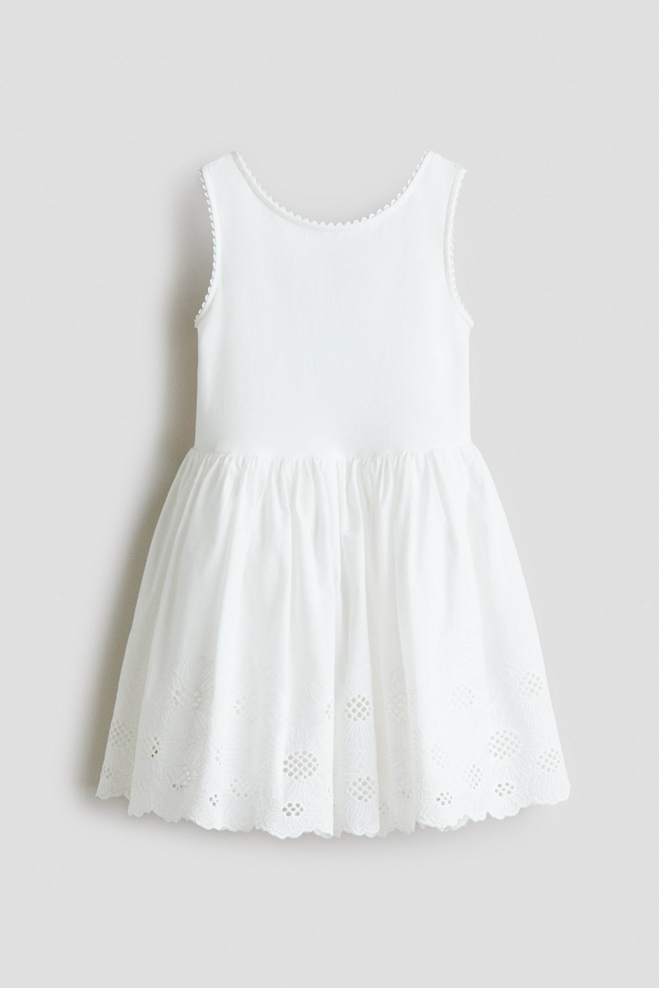 Größeres Bild ansehen: Ein weißes ärmelloses Kleid zeigt seine Vorderansicht und verfügt über ein anliegendes Oberteil mit Muschelsaum entlang des Ausschnitts und der Armlöcher, das in einen gerafften Rock übergeht. Der Rock zeigt Lochstickereimuster über einem Muschelsaum.