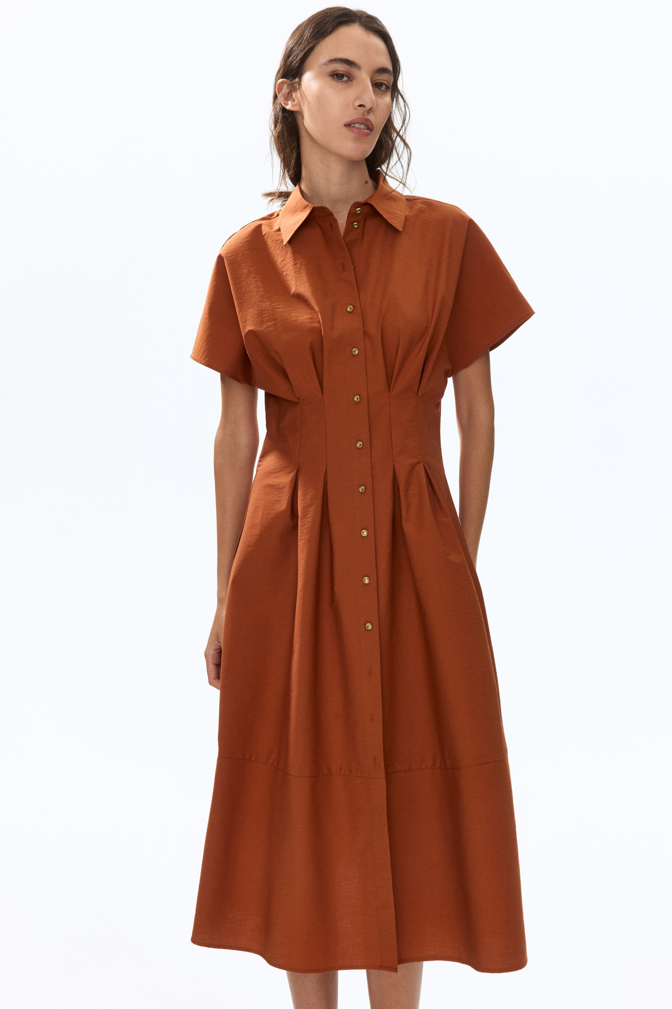 View larger image: A calf-length dress in a rust-coloured crisp weave features a double-layered collar, short sleeves, and a button-down front. Sewn-in pleats shape the waist, and the model is wearing the dress with her hands resting in its side pockets.
